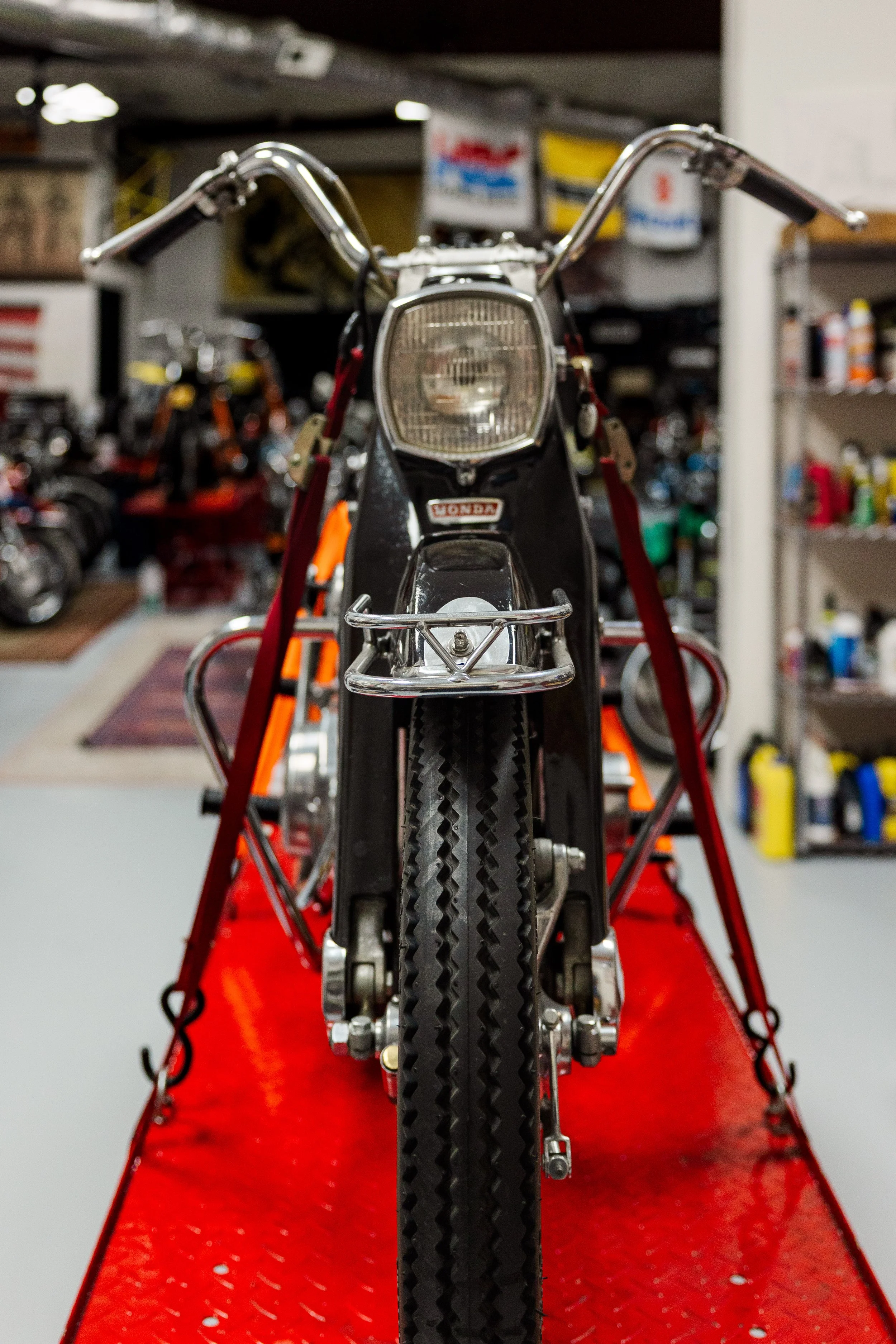 Front view of a vintage Honda motorcycle on a red display stand in a workshop or garage with shelves and supplies in the background. Vintage motorcycle detail at Birmingham Vintage Cycle, a vintage motorcycle restoration shop in Birmingham Alabama