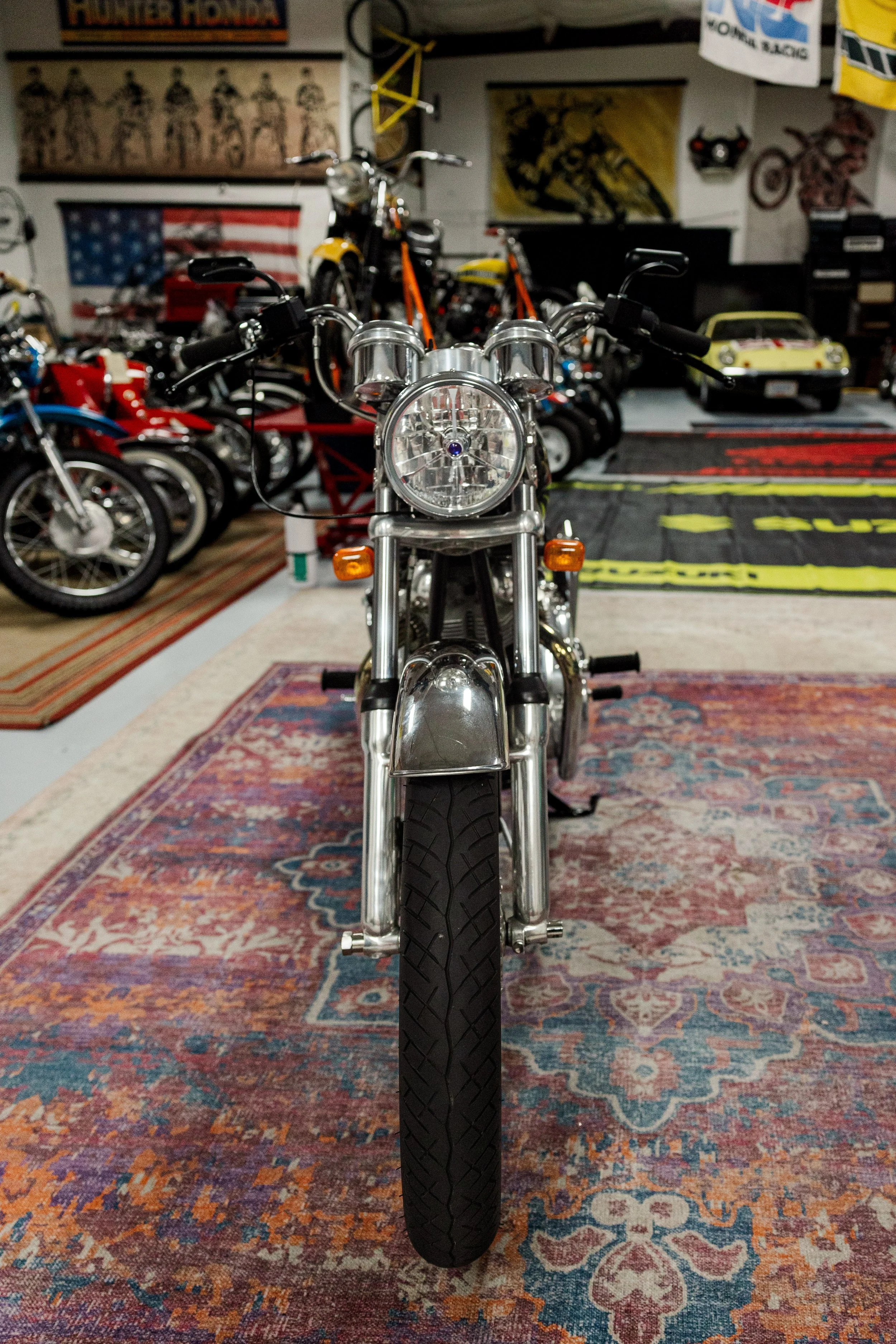 Front view of a motorcycle in a room with various other bikes and posters on the walls, including an American flag and racing posters, with patterned rugs on the floor. Vintage motorcycle detail at Birmingham Vintage Cycle, a vintage motorcycle 