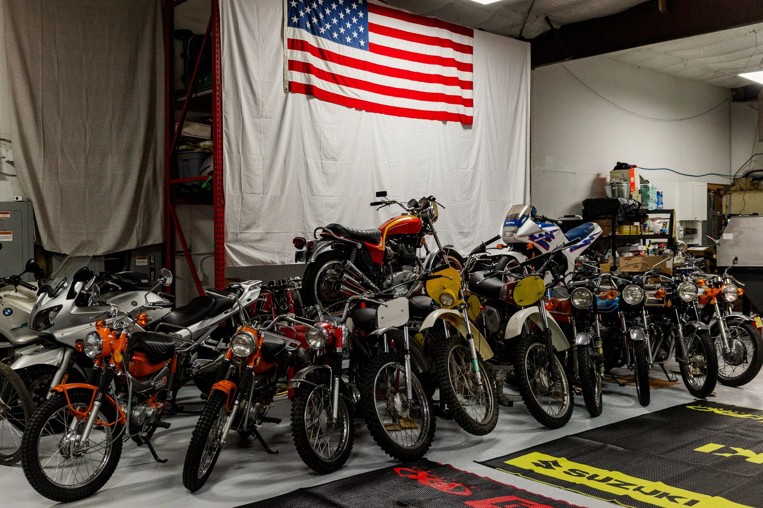 A garage with a large American flag hanging on white fabric backdrop on the wall. In front, there are multiple motorcycles parked in a row, including Honda and BMW models, with some other small bikes and scooters. Vintage motorcycle collection 