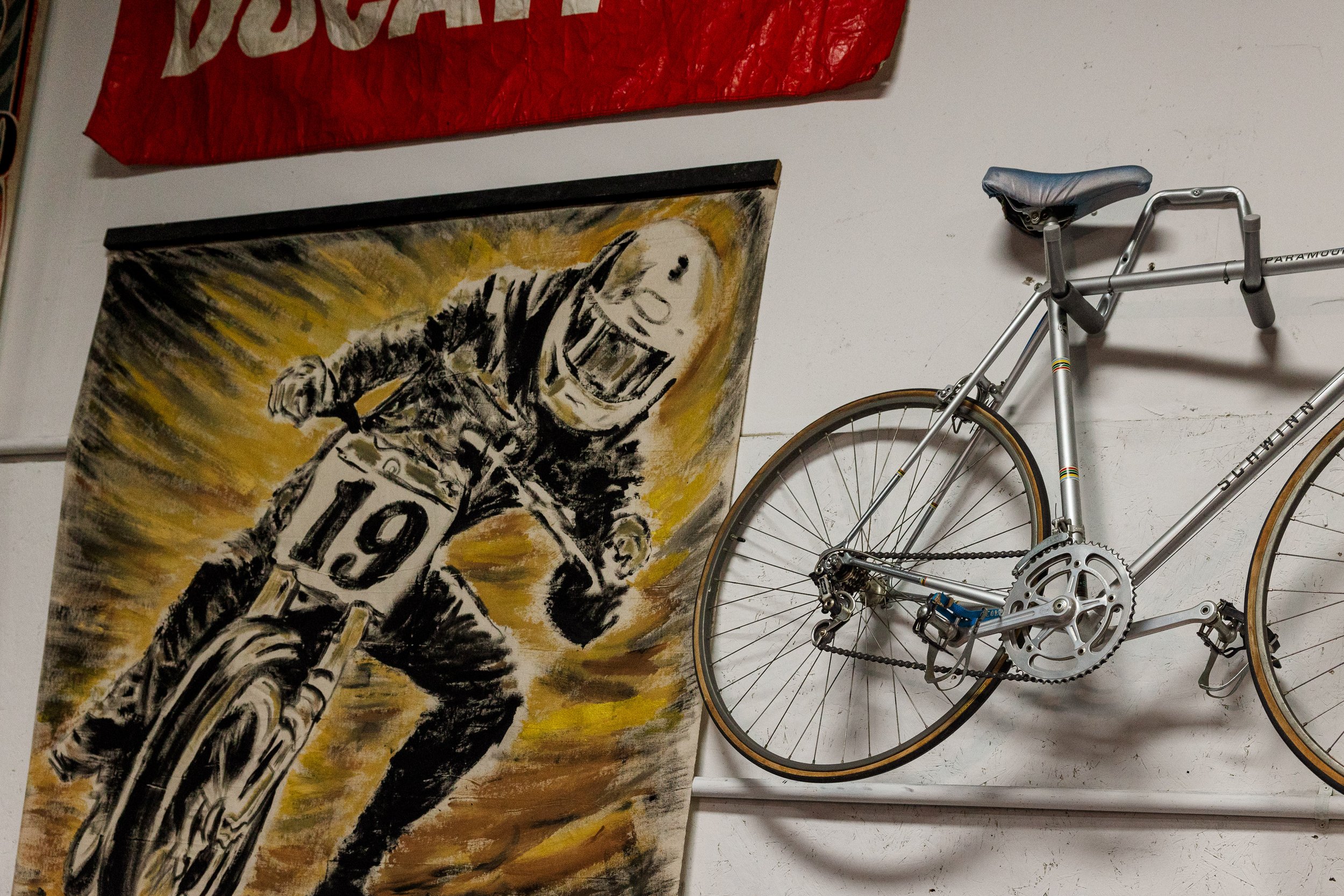 An indoor space featuring a painting of a cyclist, a red banner, and a silver bicycle mounted on the wall. Vintage motorcycle collection on display at Birmingham Vintage Cycle in Birmingham Alabama