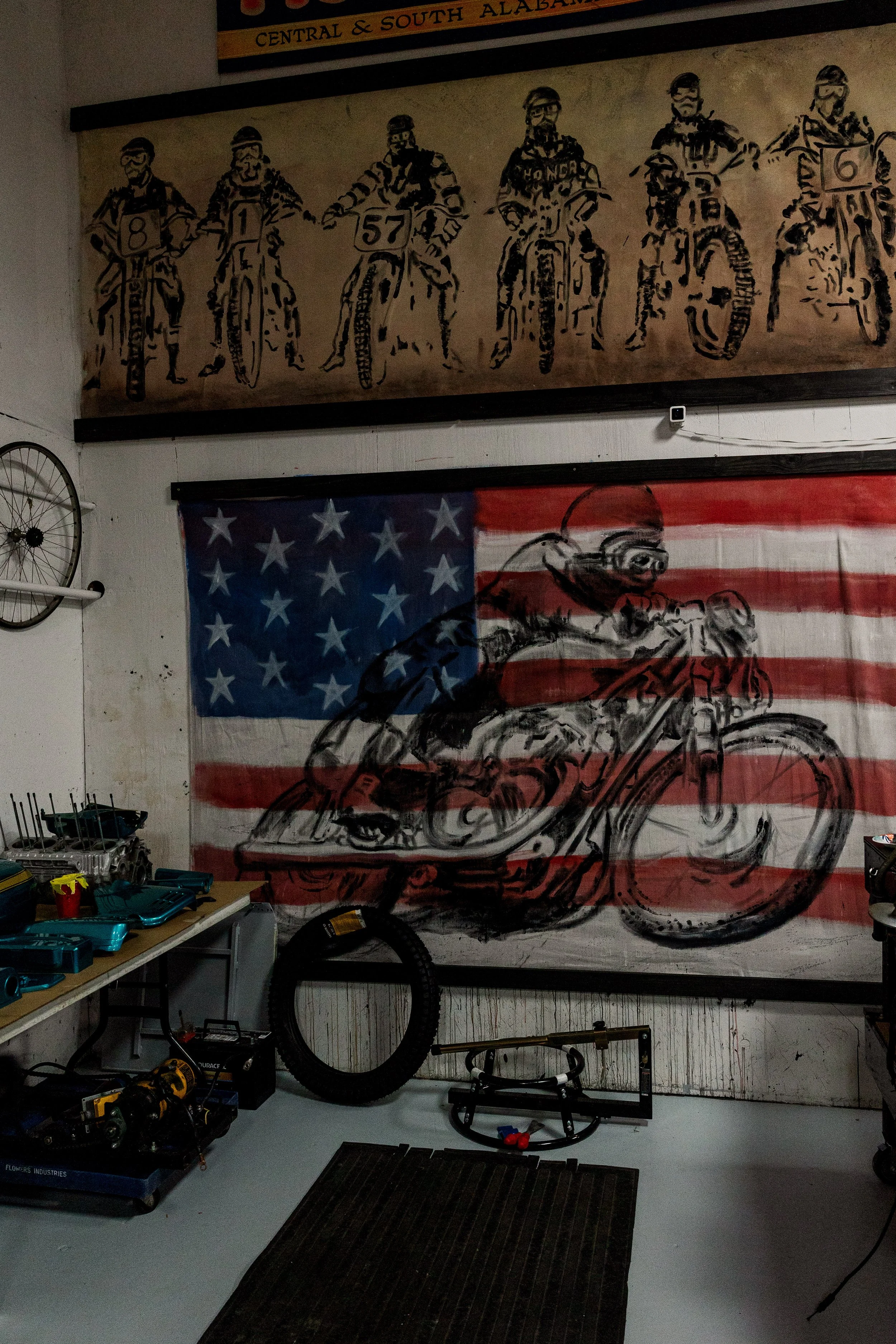 Workshop with tools organized on a workbench, a large motorcycle mural with an American flag background, and a wall hanging with a sketch of six racers and a banner reading 'Central & South Alabama'. Vintage motorcycle collection on display