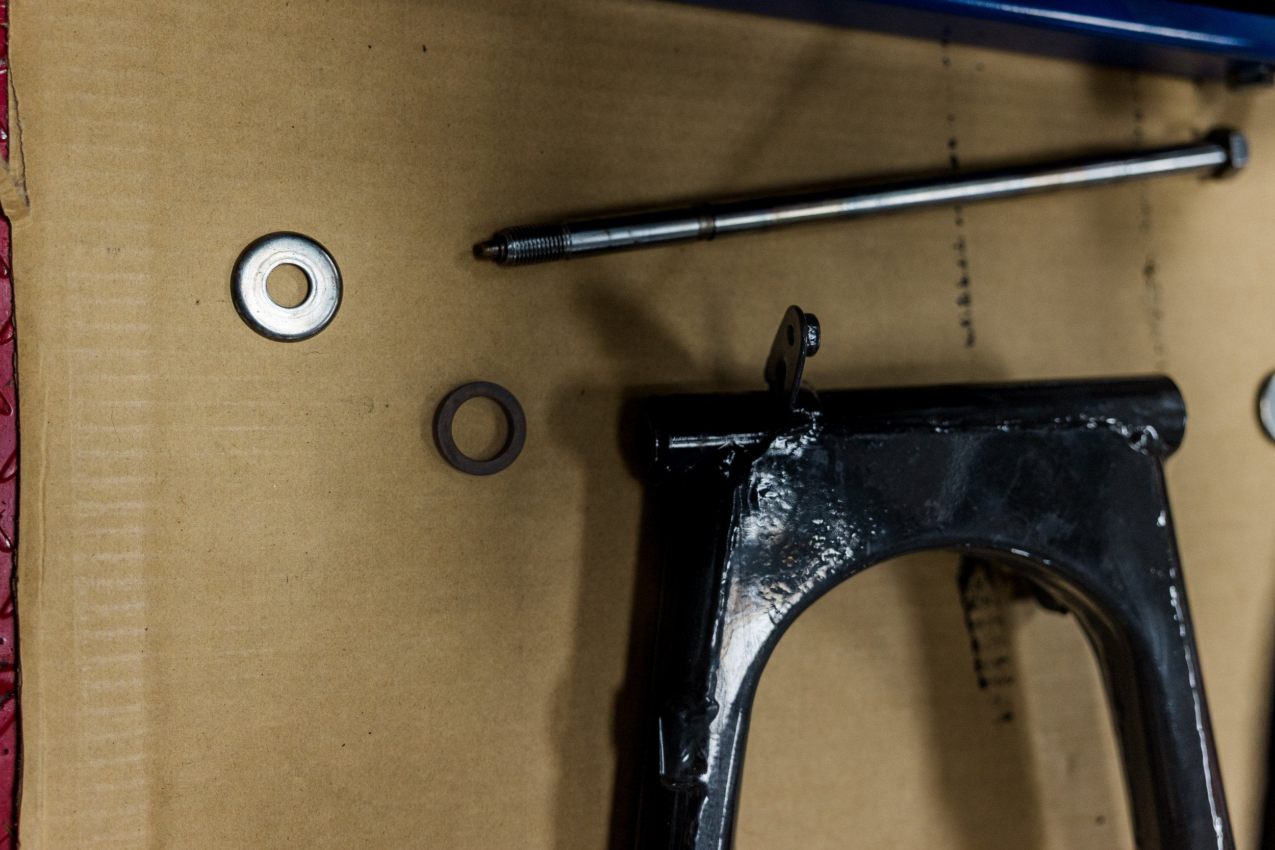 A close-up of a metal tool, possibly a part of a machine or vehicle, lying on a piece of cardboard or wood. There are washers and a black component nearby. Inside Birmingham Vintage Cycle, a vintage motorcycle shop and community space in Birmingham 