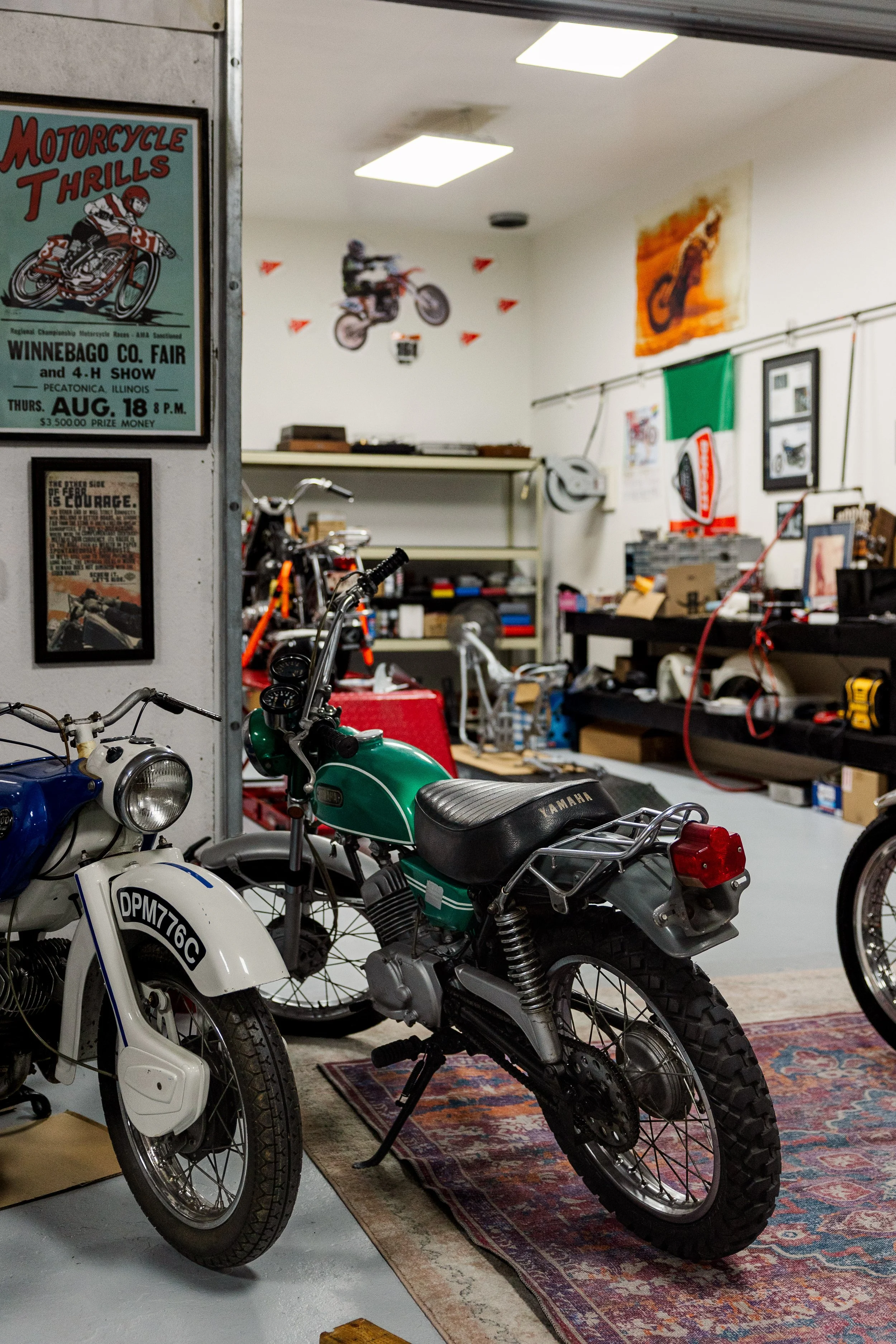 A room with vintage motorcycles and motorcycle memorabilia. In the foreground, there are white and green motorcycles with black seats. The wall on the left has a poster for a motorcycle thrill event, and the back wall displays various pictures, flags