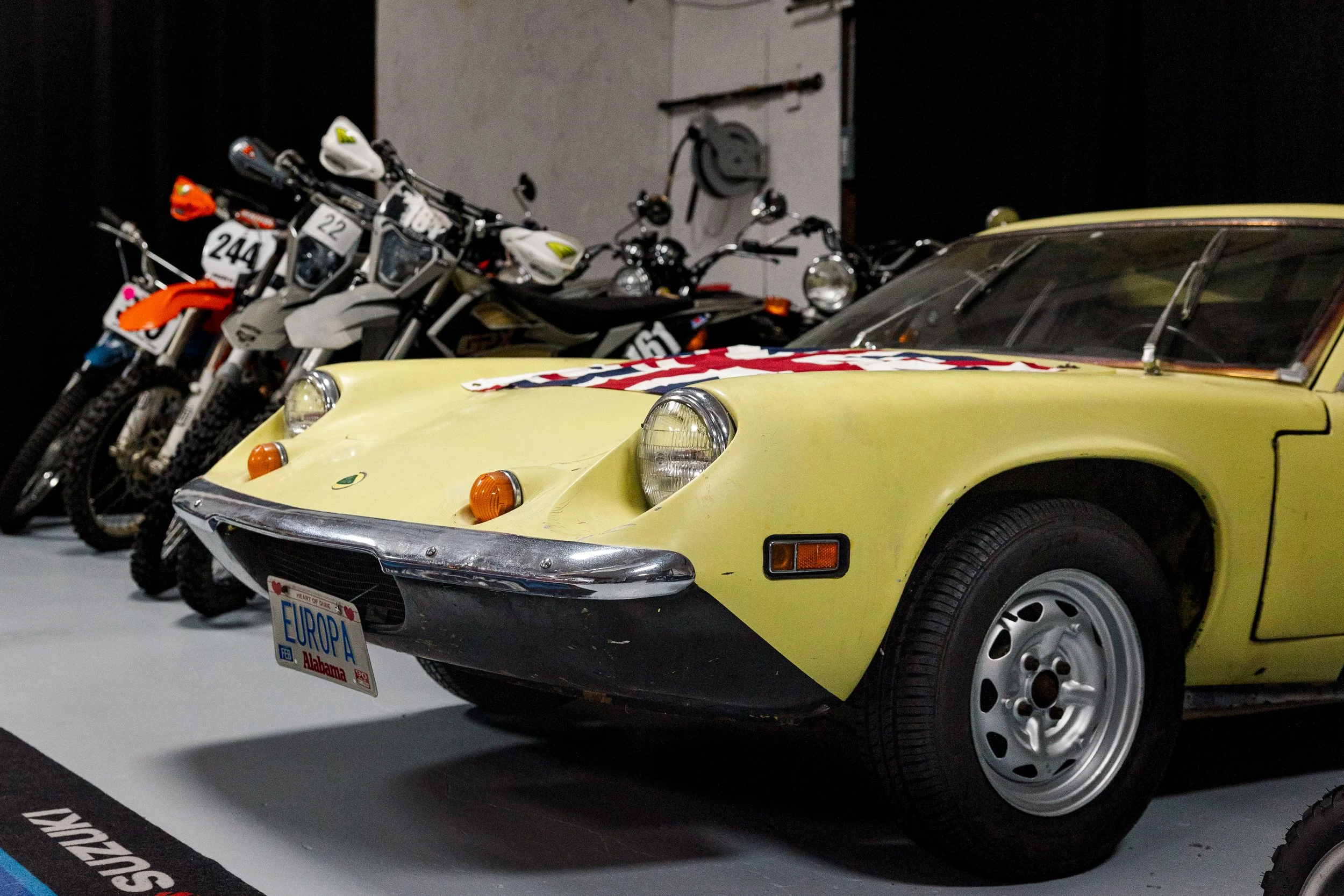 A yellow vintage sports car with a U.S. flag-themed scarf on the hood, parked next to several motocross bikes in an indoor exhibition. Vintage motorcycle detail at Birmingham Vintage Cycle, a vintage motorcycle restoration shop in Birmingham Alabama