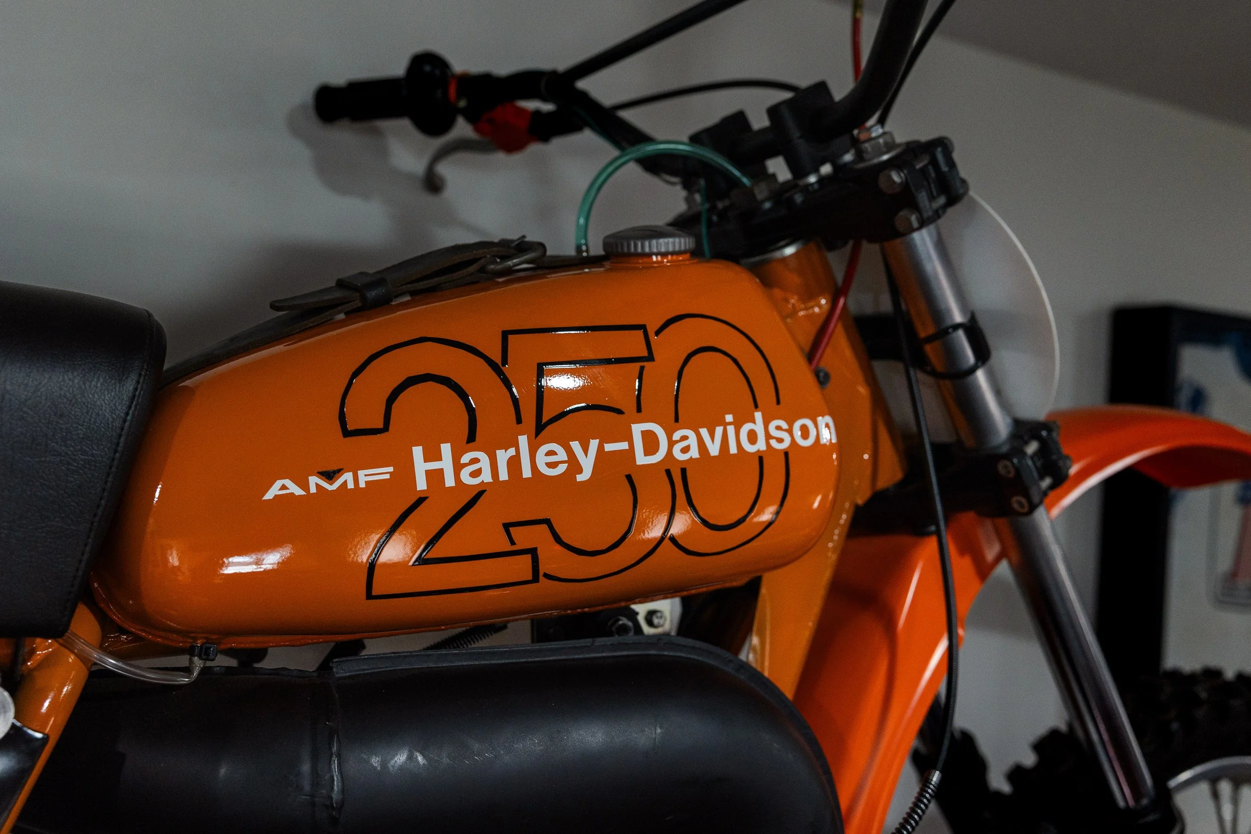Close-up of an orange Harley-Davidson motorcycle, showing part of the fuel tank with black text and logo, and the front suspension. Vintage motorcycle collection on display at Birmingham Vintage Cycle in Birmingham Alabama