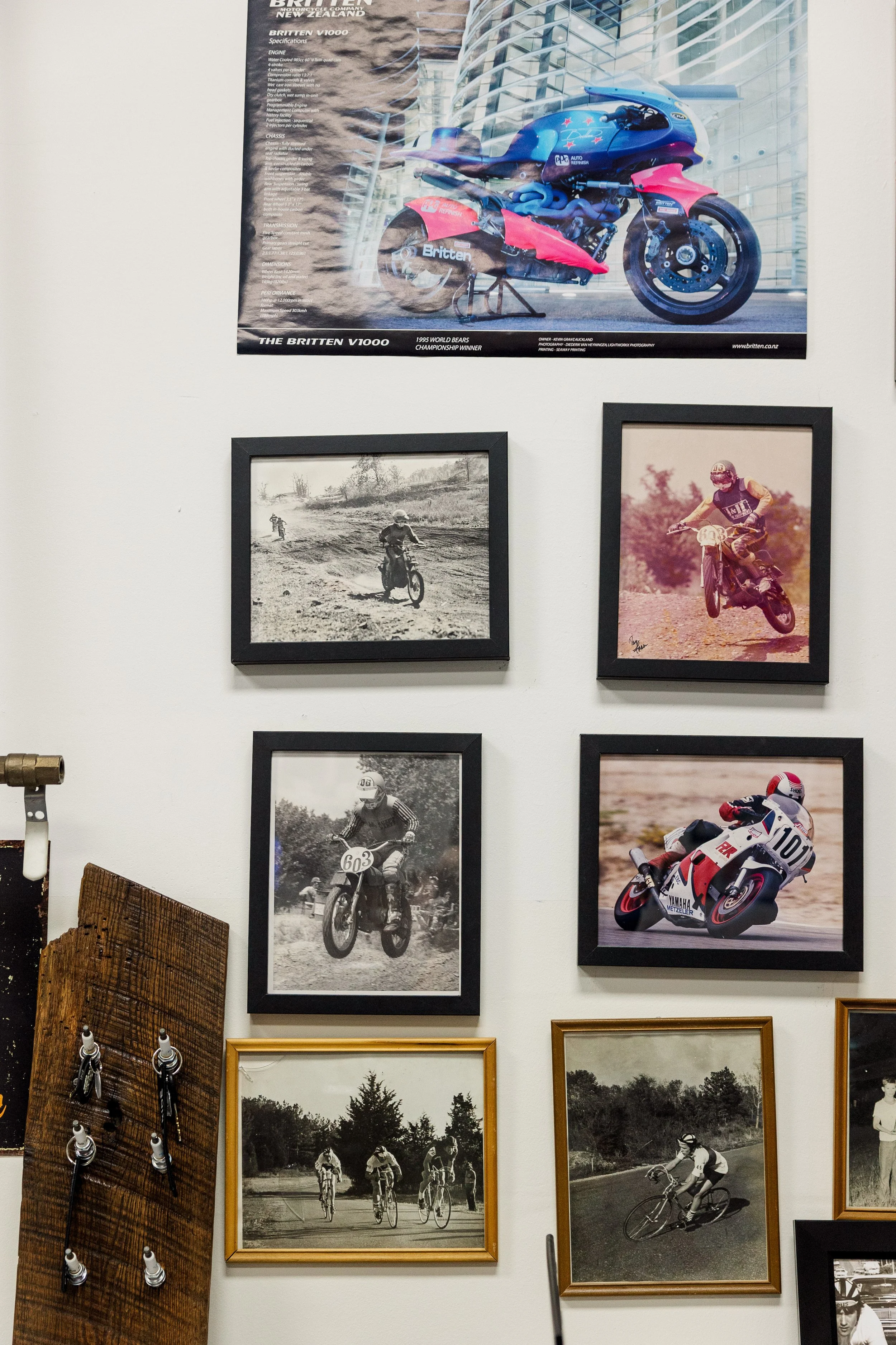 A framed poster of a blue and pink race motorcycle with text about specifications, mounted on a white wall above black-framed photographs of motorcycle and cycling scenes, including dirt biking, motocross, and road cycling.