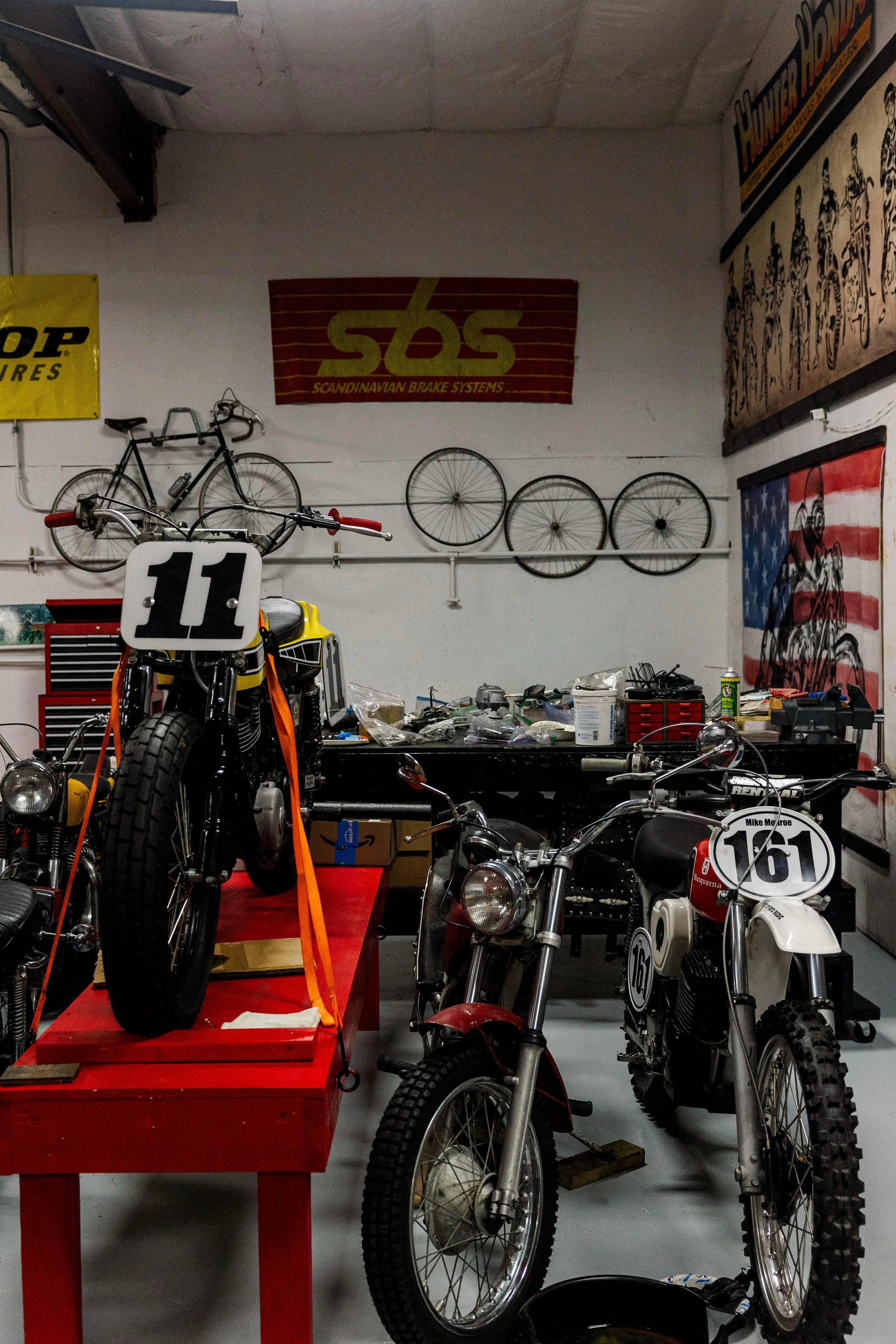 A garage or workshop with vintage motorcycles, bike wheels mounted on the wall, and various tools and supplies on the workbench. Vintage motorcycle collection on display at Birmingham Vintage Cycle in Birmingham Alabama