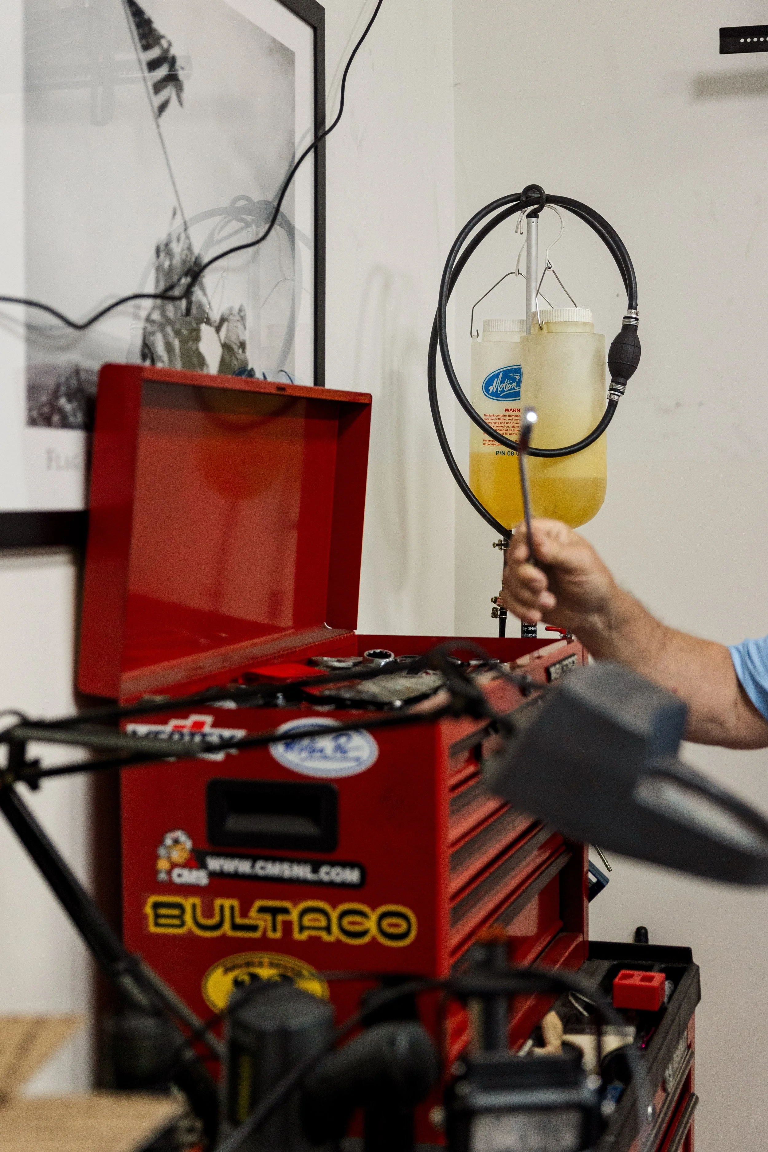 A red tool chest labeled 'Bulaco' with various tools on top, a yellow fluid container hanging on the wall, and a hand holding a tool near the chest in a workshop or garage setting. Inside Birmingham Vintage Cycle, a vintage motorcycle shop 