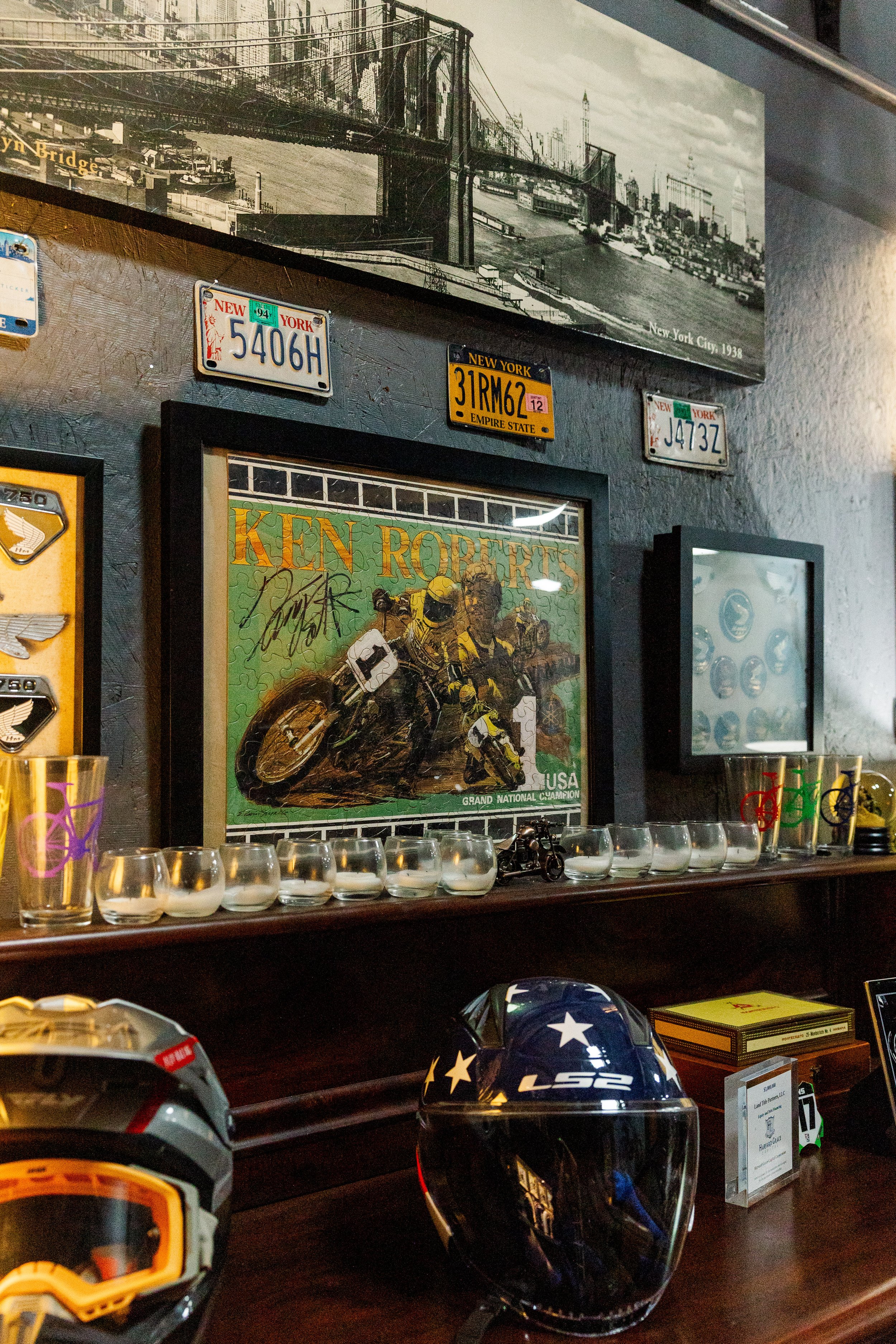 Decorative items on a wooden surface, including a motorcycle helmet with stars, glasses with star designs, and a framed poster of a motorcycle race. In the background, there are various vintage license plates from New York and a black-and-white New Y