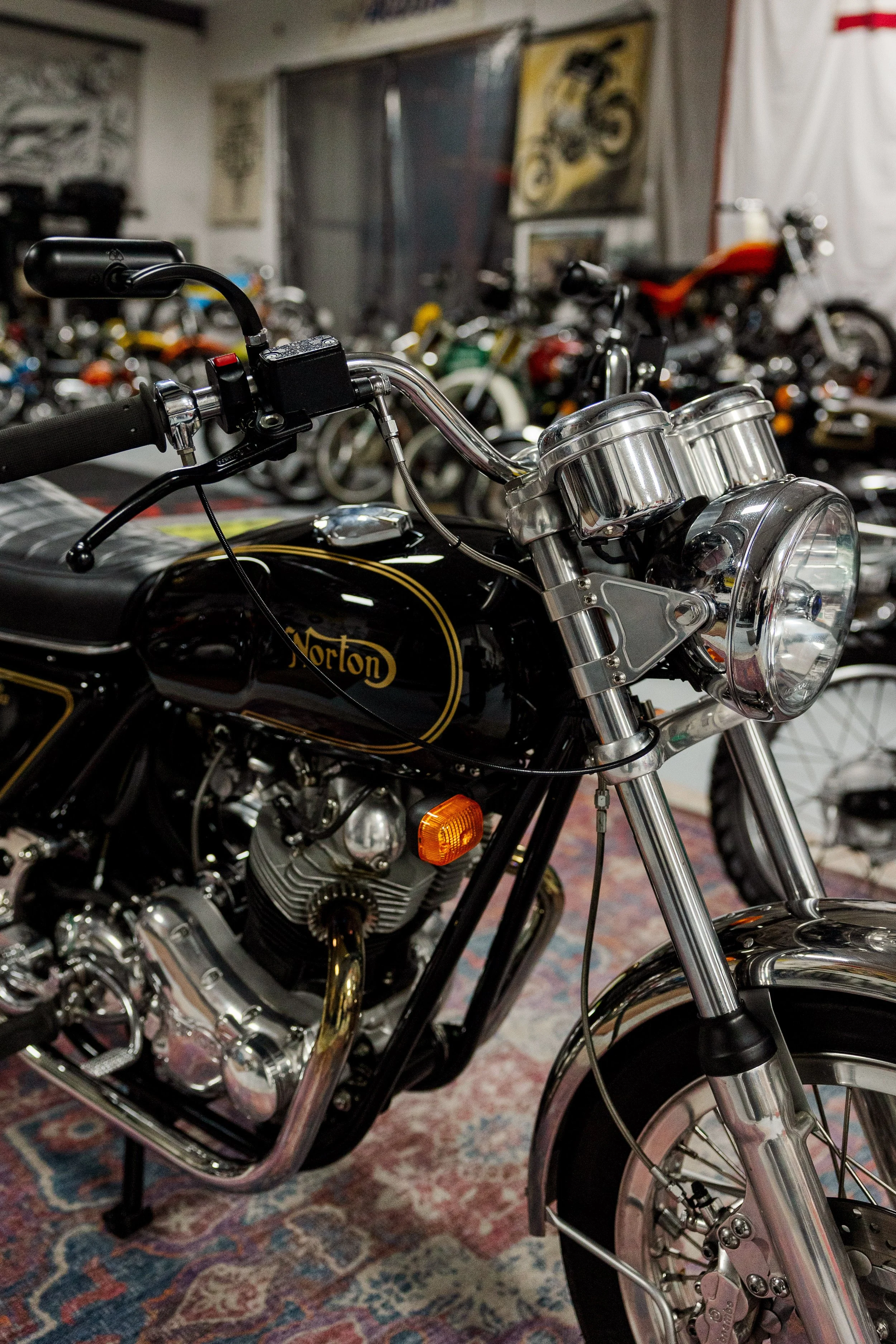 Close-up of a vintage Norton motorcycle with chrome detailing, black fuel tank, and multiple bikes in the background in a showroom or garage setting. Vintage motorcycle detail at Birmingham Vintage Cycle, a vintage motorcycle restoration shop in