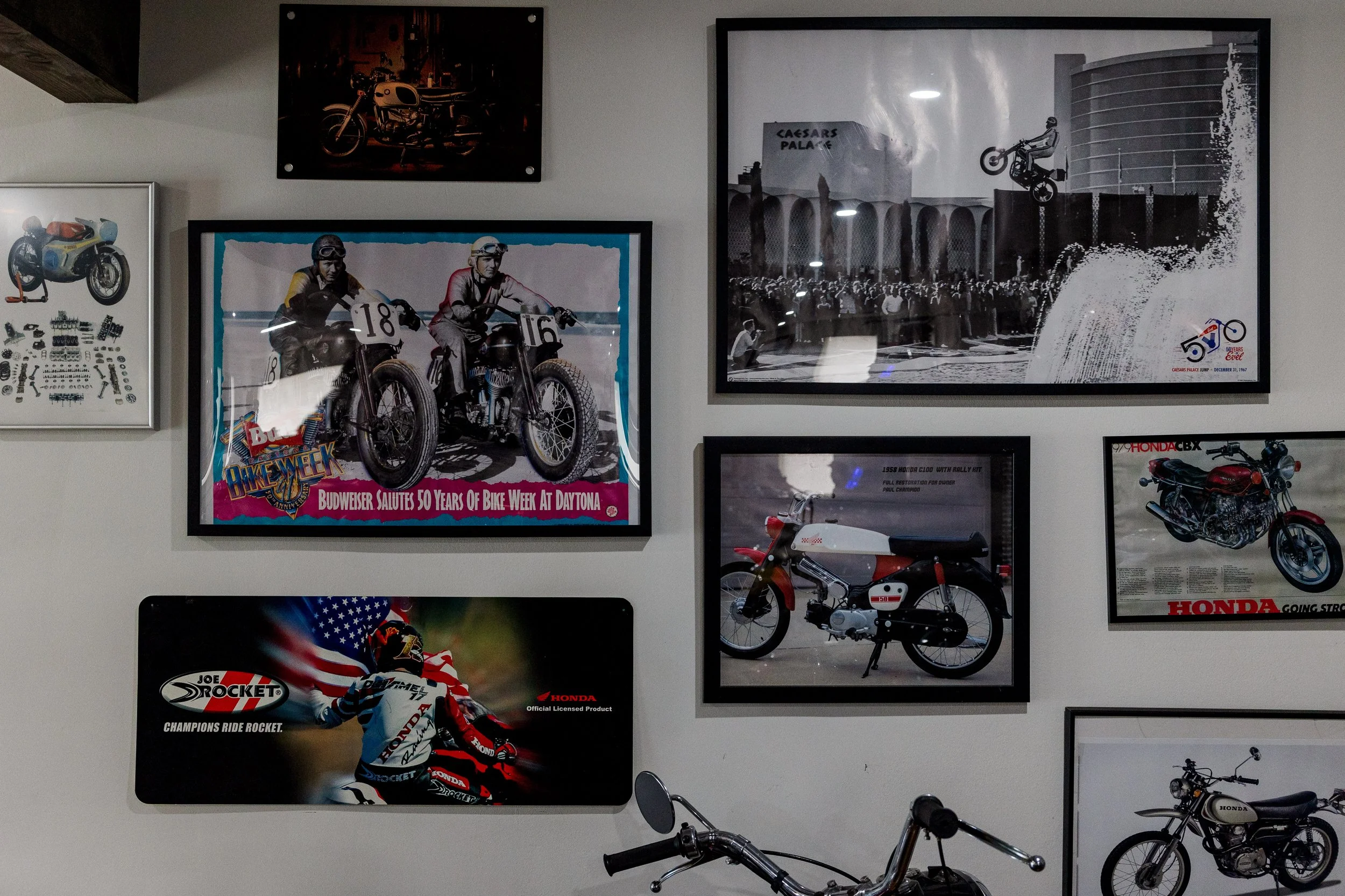 Collection of vintage motorcycle posters and photographs displayed on a wall, including images of racers, motorcycles, and related memorabilia. Vintage motorcycle collection on display at Birmingham Vintage Cycle in Birmingham Alabama