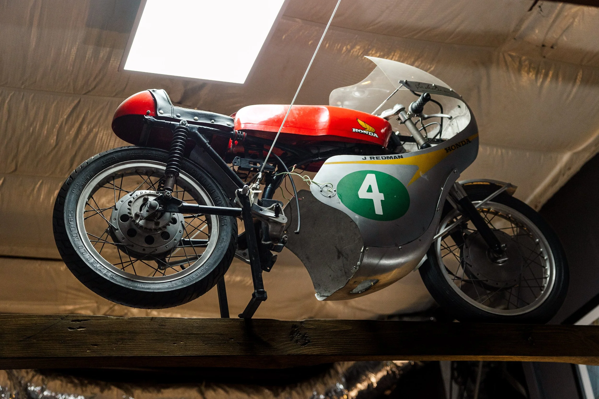 A vintage racing motorcycle displayed on a wooden platform in a museum, featuring a red fuel tank with the Honda logo, a large green number 4 on the side, and a partial view of the front wheel and suspension. Restored vintage motorcycle in BVC
