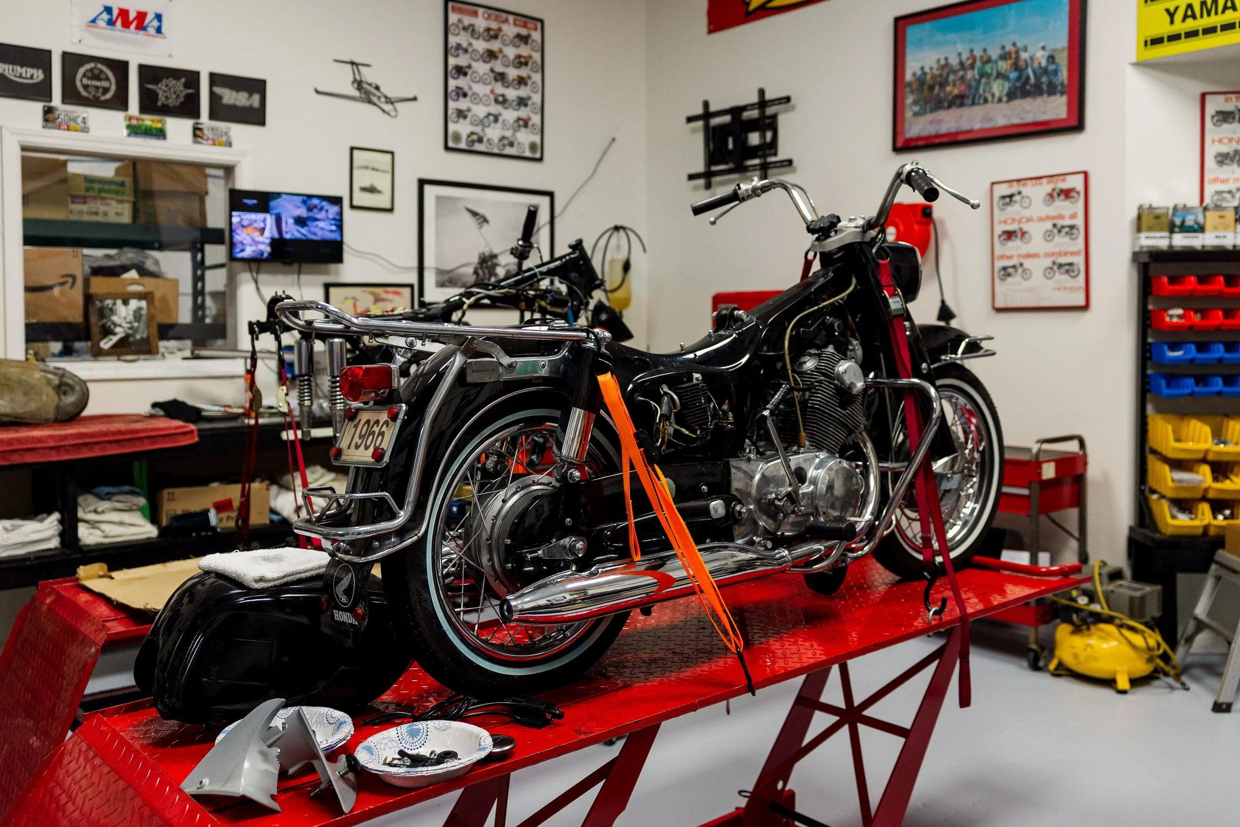 A vintage black motorcycle with a chrome frame on a red work table in a workshop or garage surrounded by motorcycle parts and tools. The background includes framed pictures, posters, and shelves with boxes and equipment.