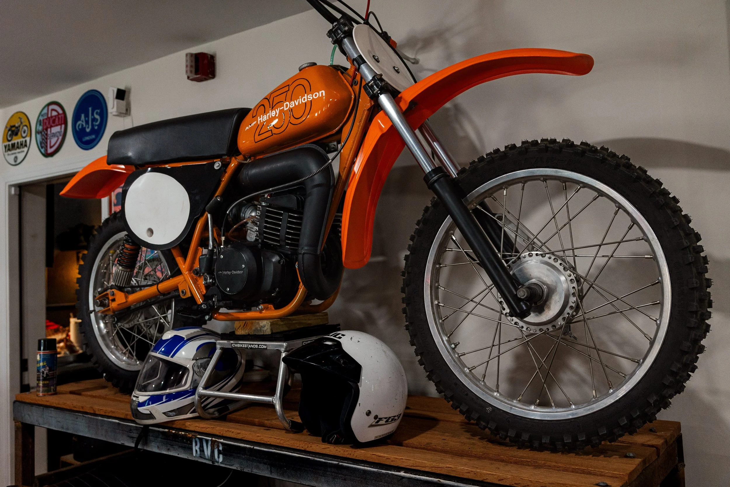 A motorcycle with orange and black parts displayed on a wooden shelf, with racing helmet and motorcycle helmet below, in a room with vintage motorcycle signs on the wall. Vintage motorcycle collection on display at Birmingham Vintage Cycle