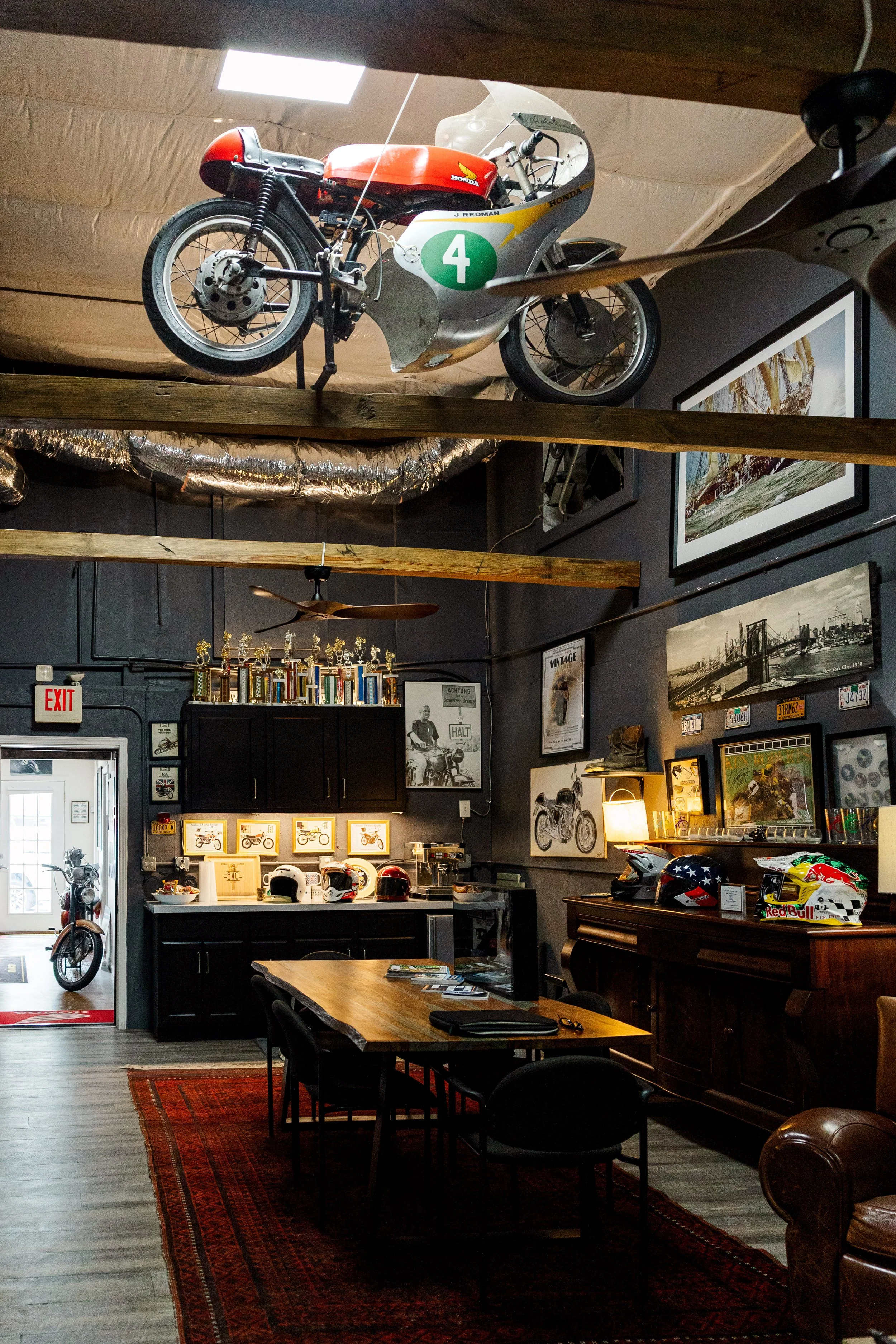Inside Birmingham Vintage Cycle, a vintage a motorcycle mounted on the ceiling, black furniture, framed pictures and posters on the dark walls, a wooden table with chairs, helmets on display, and a red and black area rug on a wooden floor.