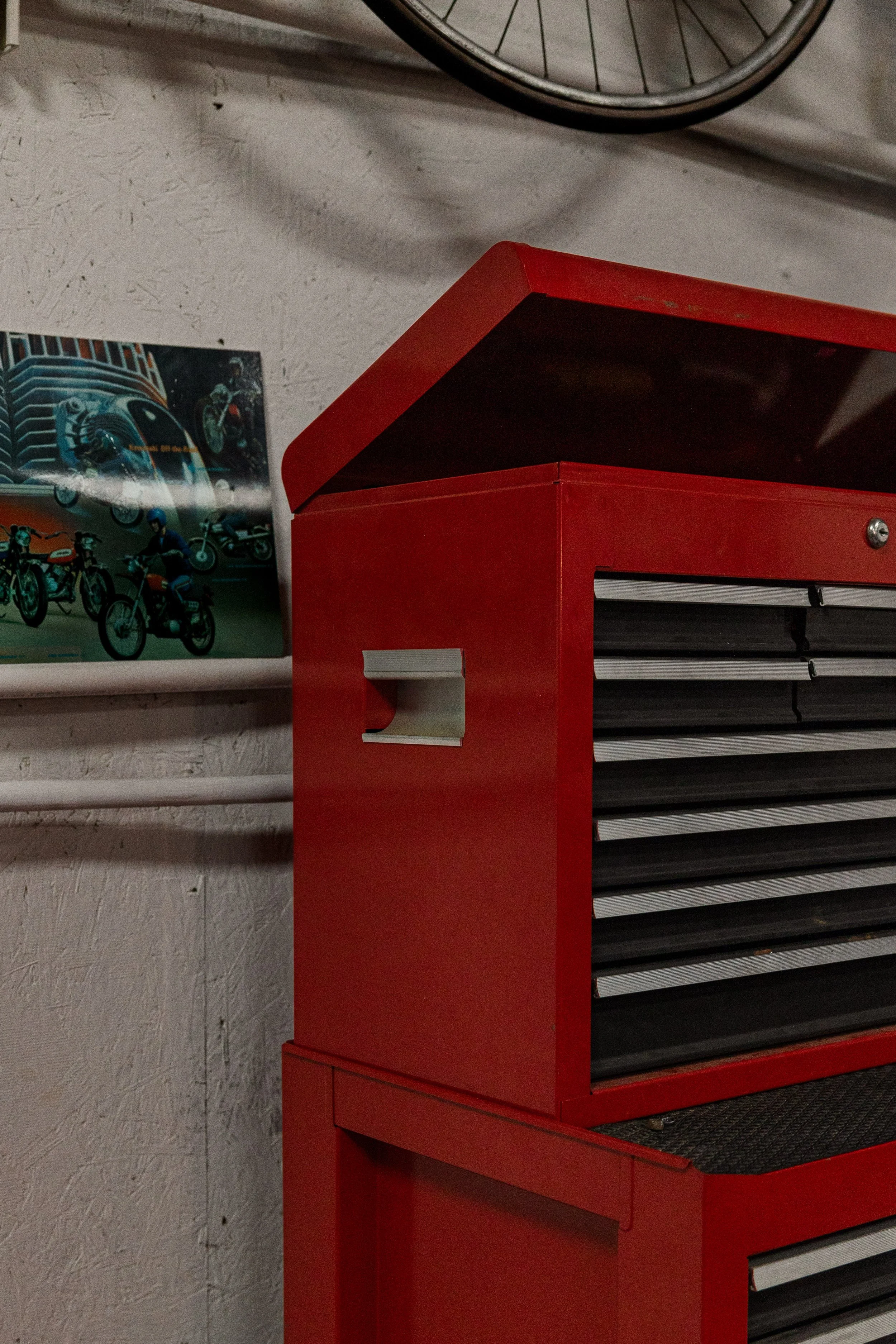 A red tool chest with black and gray drawers is placed against a textured white wall. A cartoon poster featuring motorcycles is partially visible on the wall, and a metal bicycle wheel can be seen hanging above.
