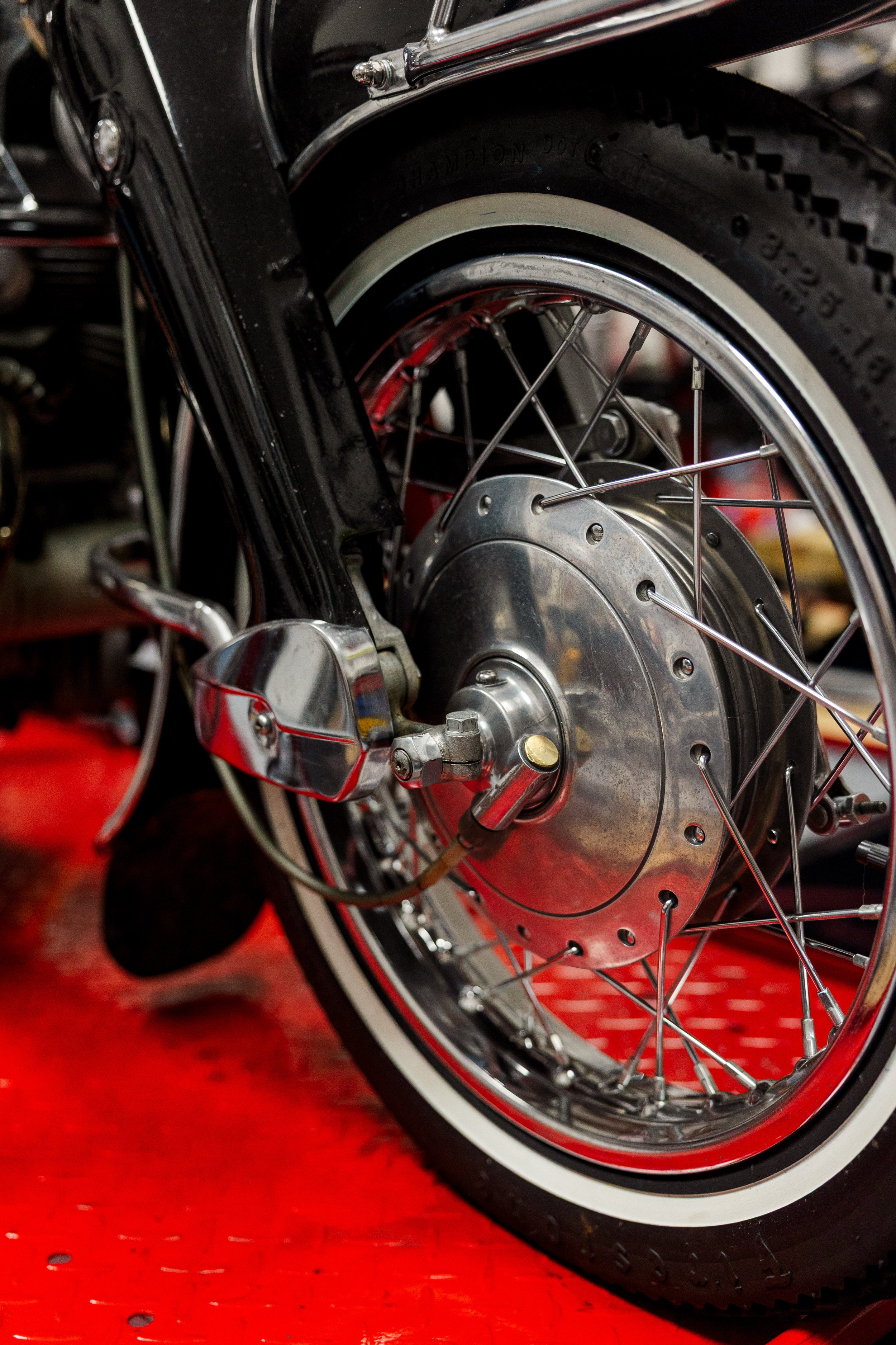 Close-up view of a motorcycle wheel with a black tire, silver spokes, and a silver hub against a red floor. Vintage motorcycle detail at Birmingham Vintage Cycle, a vintage motorcycle restoration shop in Birmingham Alabama