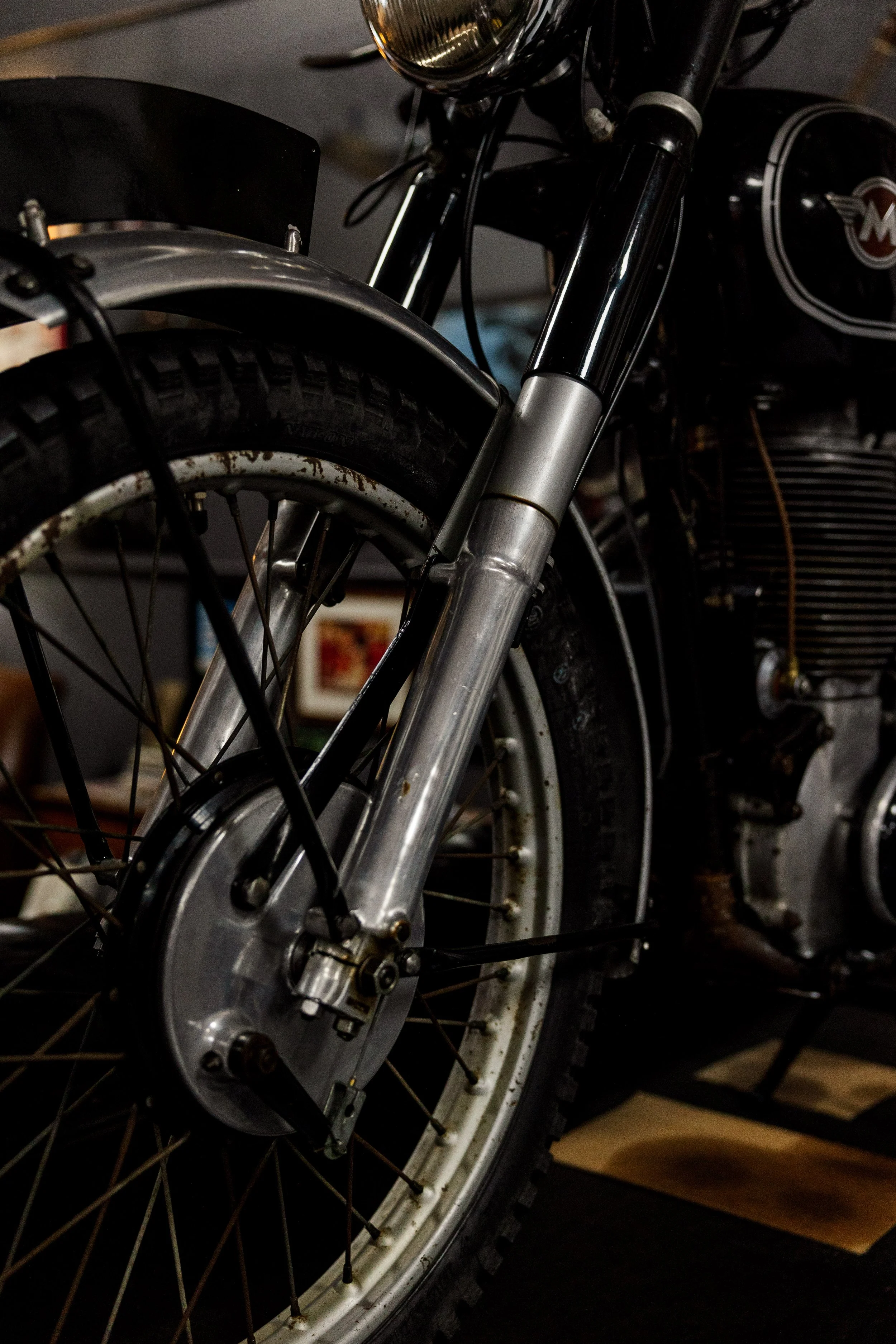 Close-up of a vintage motorcycle's front wheel, fork, and headlamp with black and silver components. The tire shows signs of tire wear and slight rust on the rim, and the motorcycle has a logo on the tank. The background is out of focus with a framed