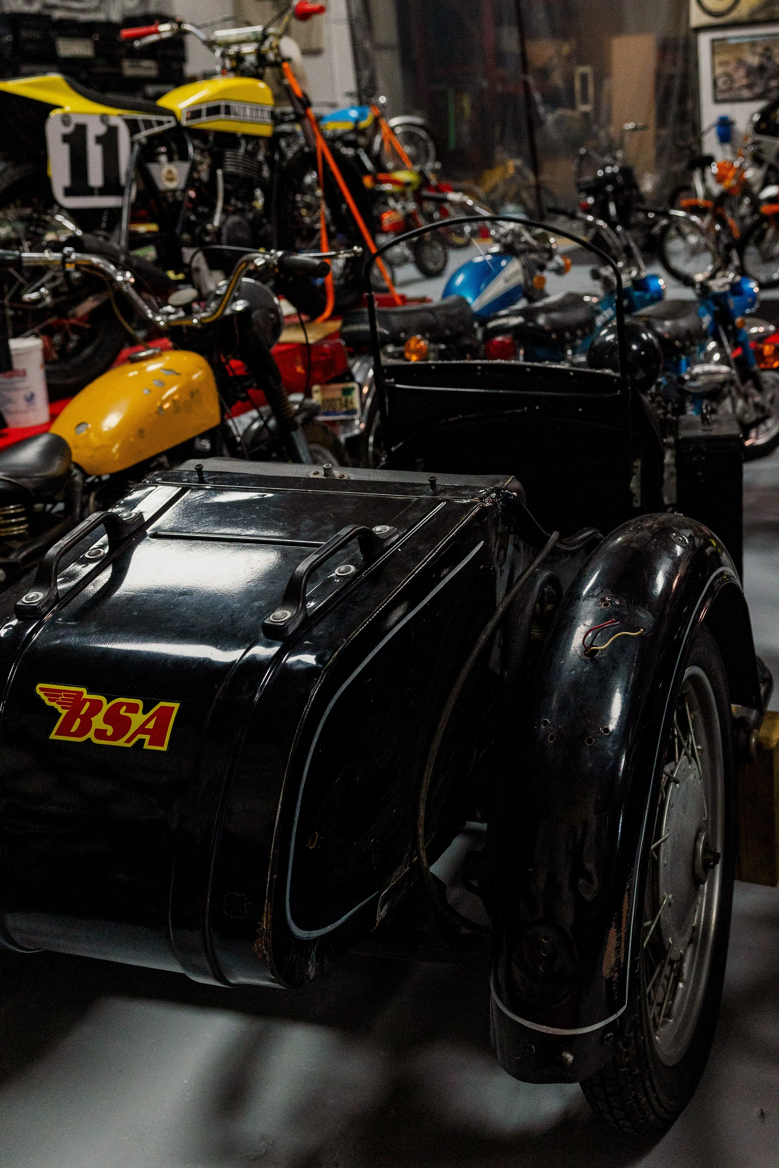 A collection of vintage motorcycles and a black sidecar displayed indoors, with some motorcycles having yellow and blue paint and others orange accents in a garage or workshop setting. Vintage motorcycle collection on display at BVC