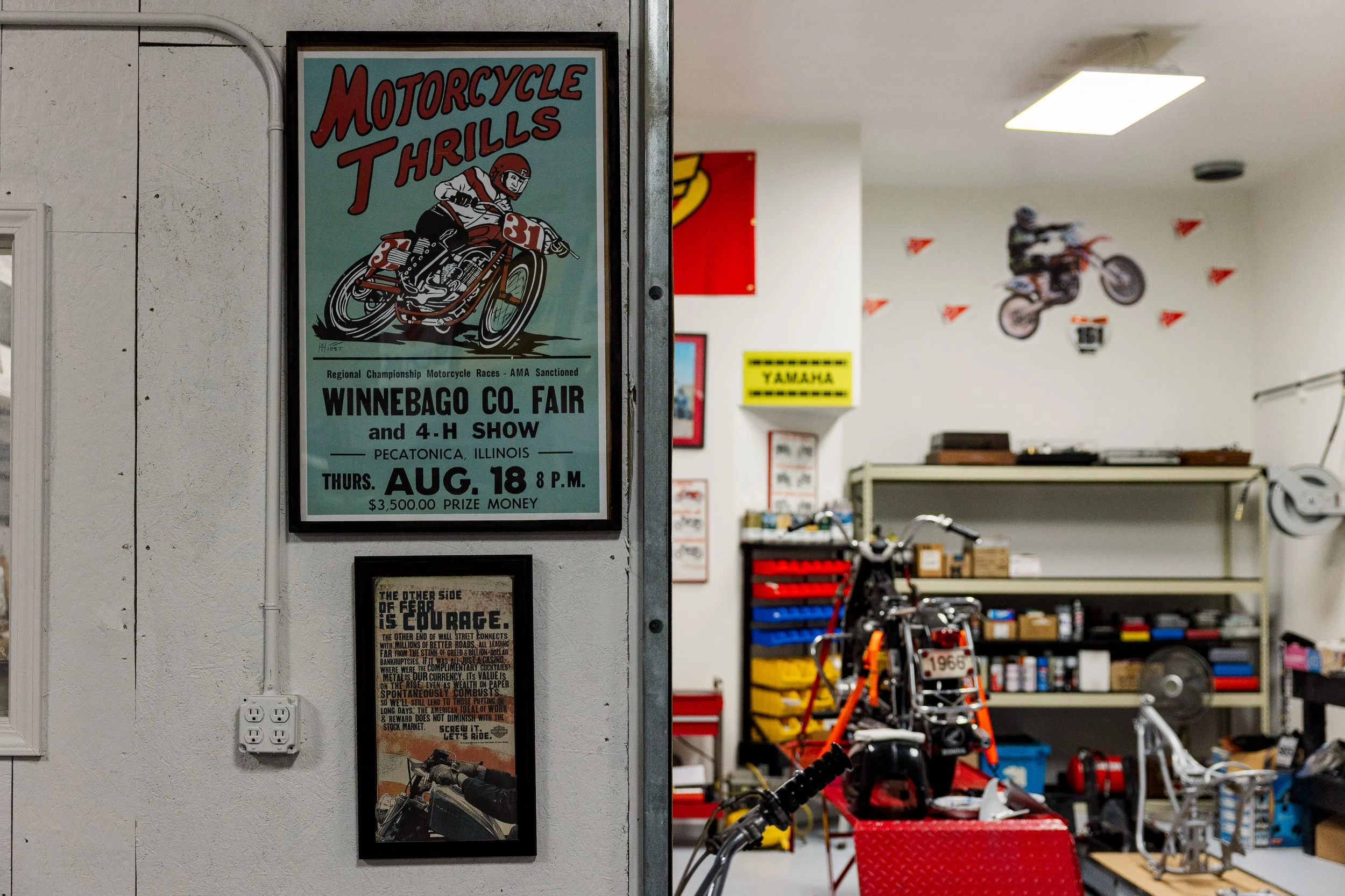 Poster advertising the 'Motorcycle Thrills' event at the Winnebago County Fair on August 18th, featuring a vintage motorcycle race illustration, located inside a workshop with bicycles, tools, and red and yellow parts visible in the background.