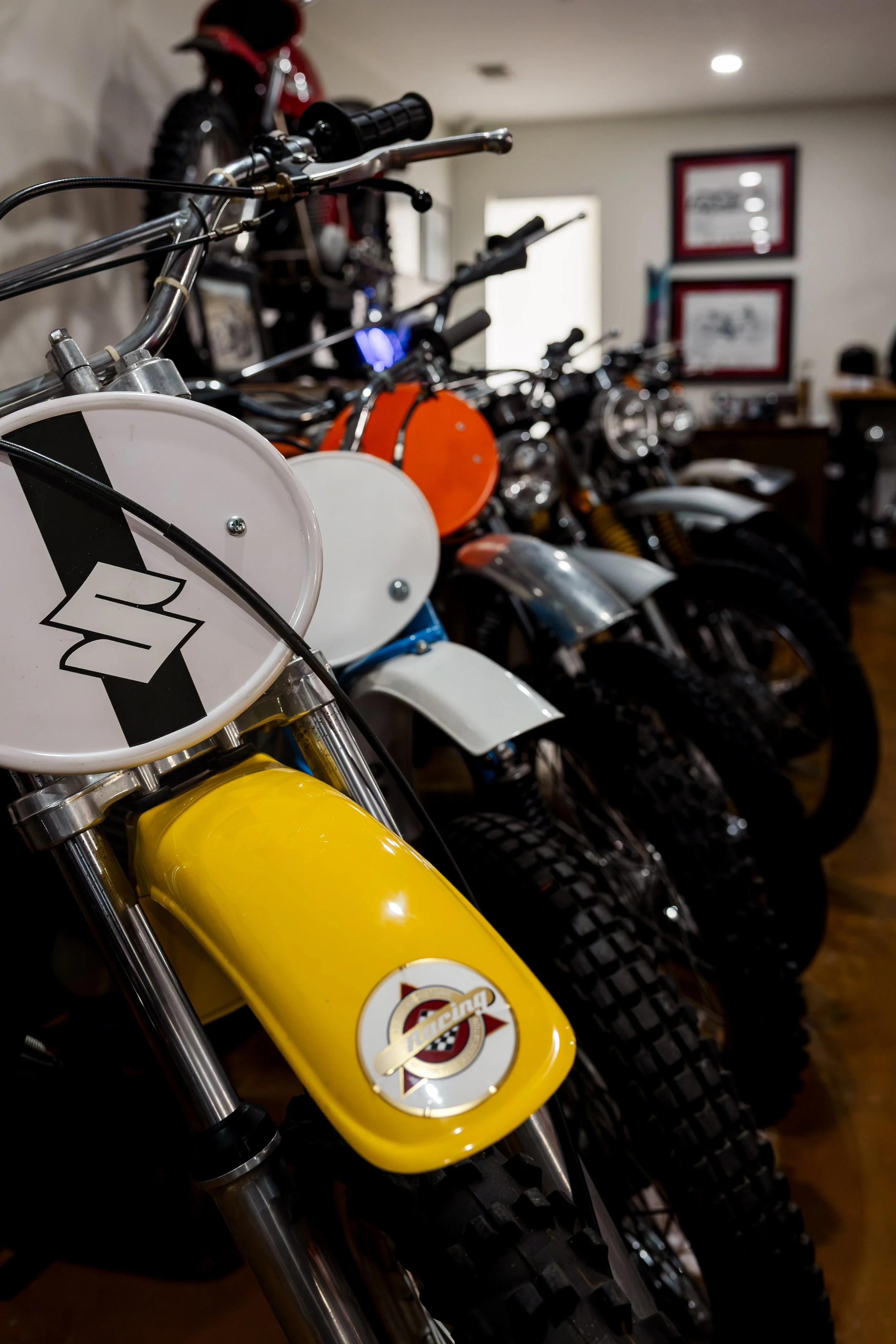 Close-up of a row of dirt bikes in a showroom, with focus on a yellow bike with a white circular number plate bearing a racing logo, and several other bikes in various colors blurred in the background. Vintage motorcycle collection on display
