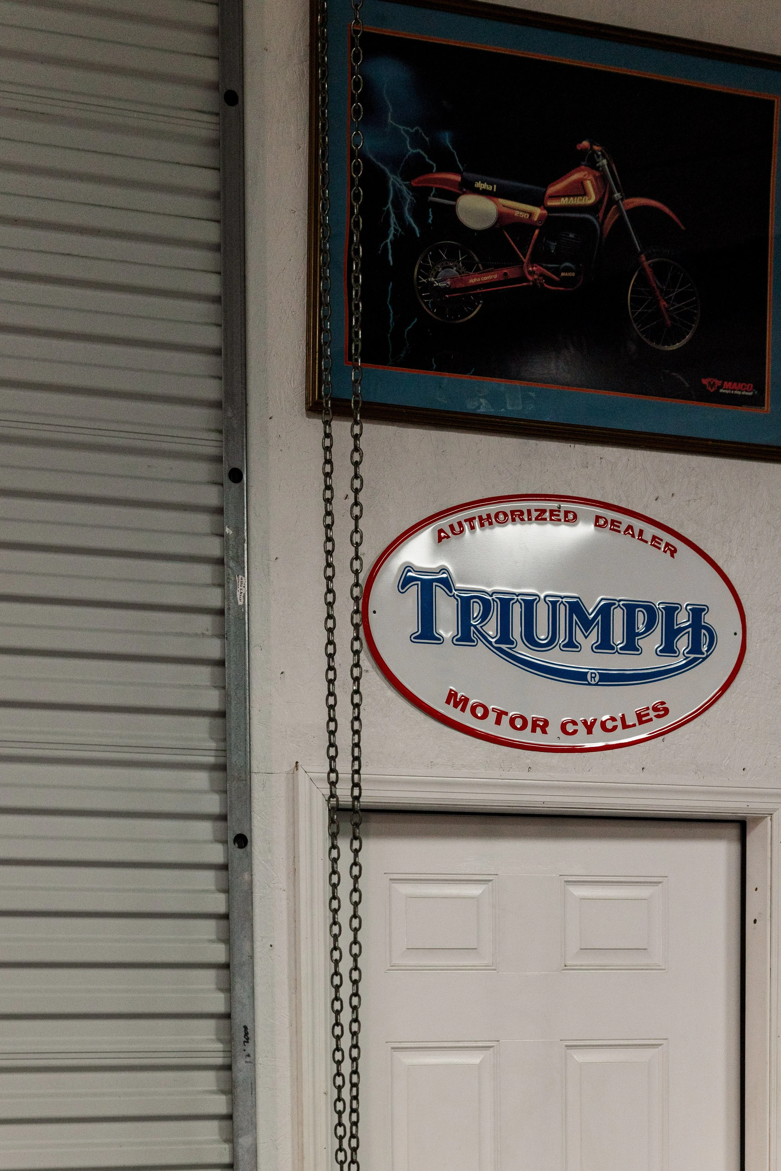 A garage with a motorcycle poster on the wall and a Triumph Motorcycles sign, with part of a garage door and a metal chain hanging. Vintage motorcycle collection on display at Birmingham Vintage Cycle in Birmingham Alabama