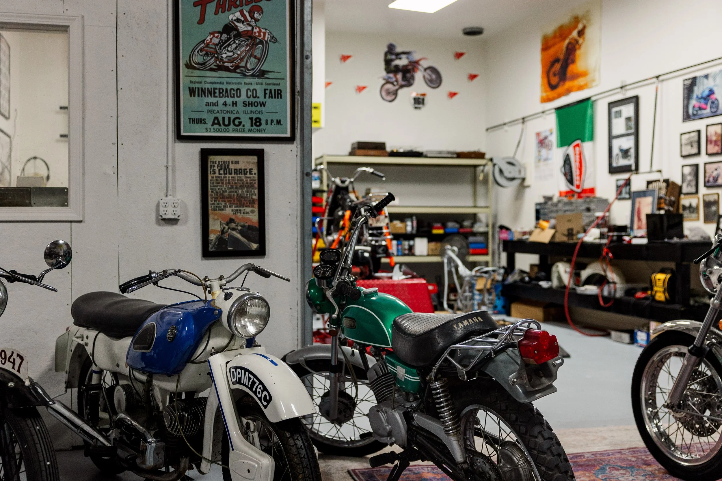 Inside a garage with vintage motorcycles and various posters on the walls, including a motorcycle race poster and other framed pictures.