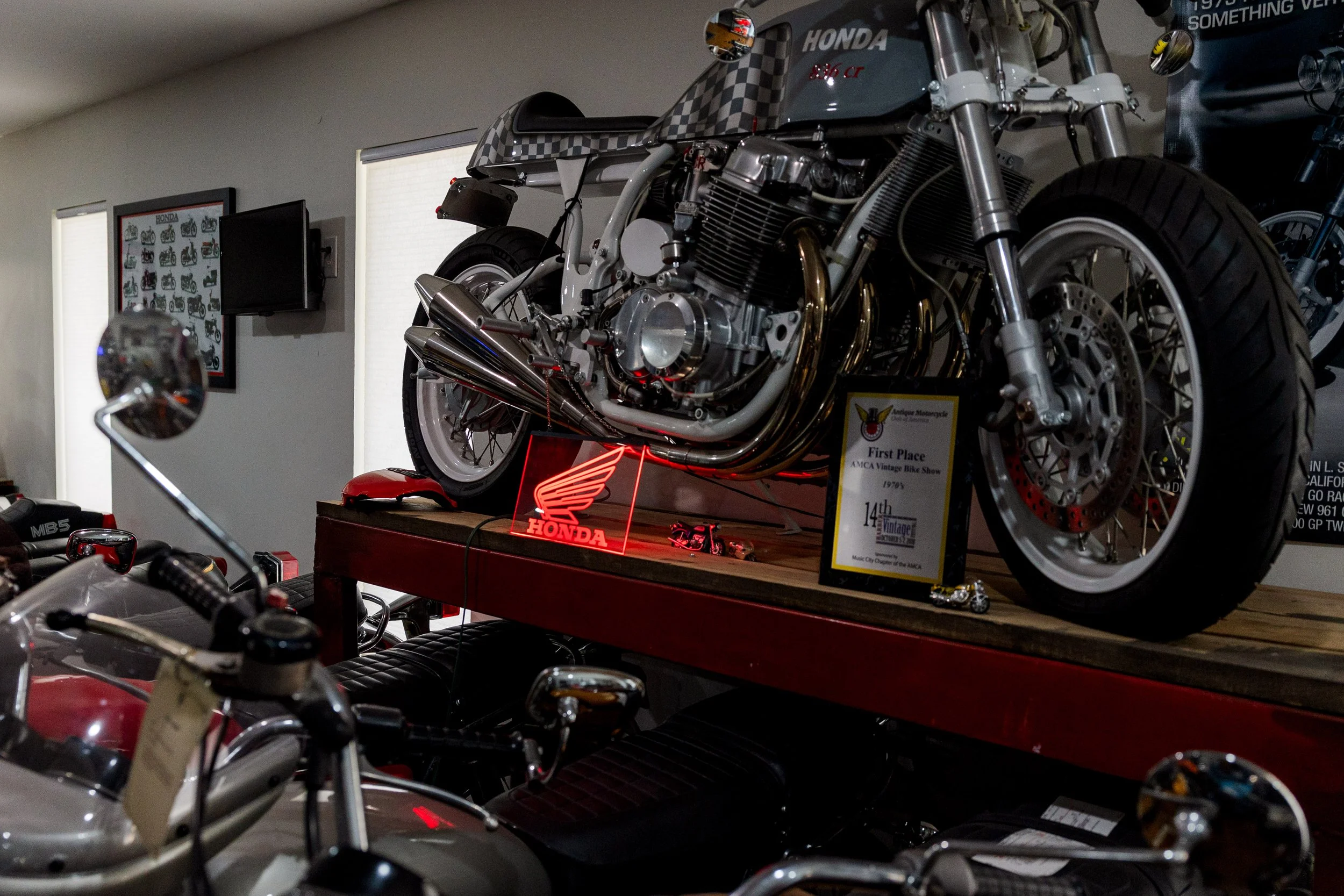 Vintage Honda motorcycle displayed on a platform at a motorcycle show, with a sign indicating first place award and collection details. Other motorcycles and memorabilia are visible in the background. Vintage motorcycle collection on display 