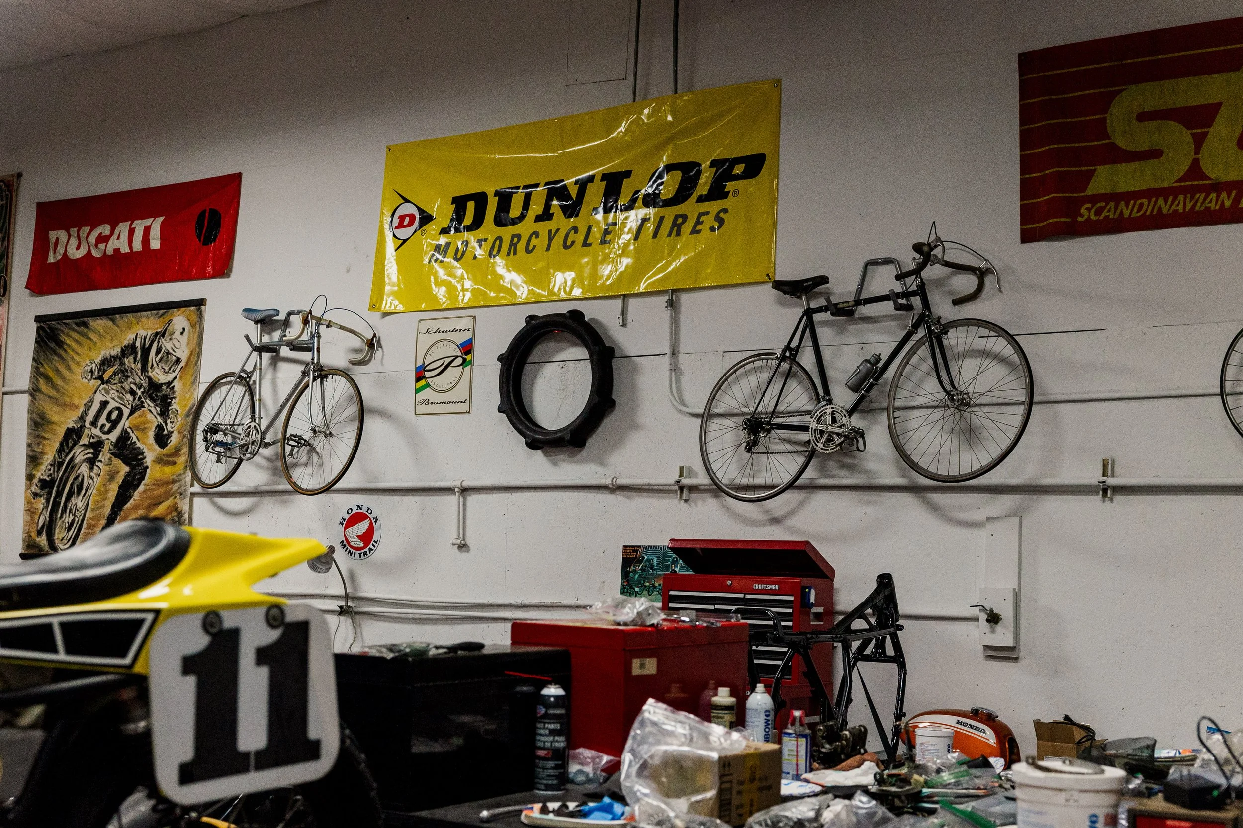 A workshop with vintage motorcycle posters, bicycles hanging on the wall, and various tools and items scattered on workbenches, including a yellow sports bike in the foreground. Vintage motorcycle collection on display at Birmingham Vintage Cycle