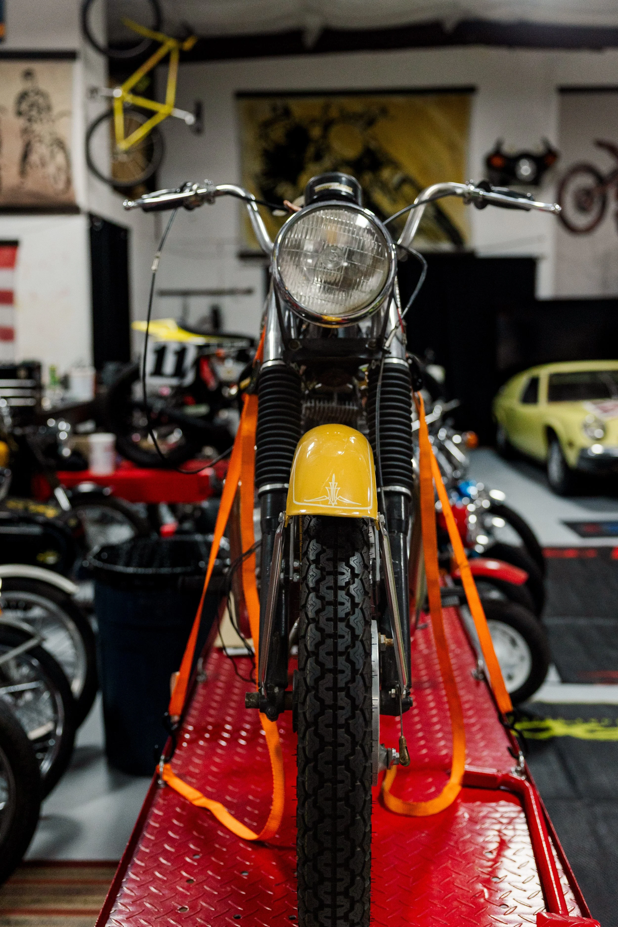 Front view of a vintage yellow motorcycle on a red transport rack inside a garage, with various bikes and wall decorations in the background. Vintage motorcycle detail at Birmingham Vintage Cycle, a vintage motorcycle restoration shop in Birmingham 