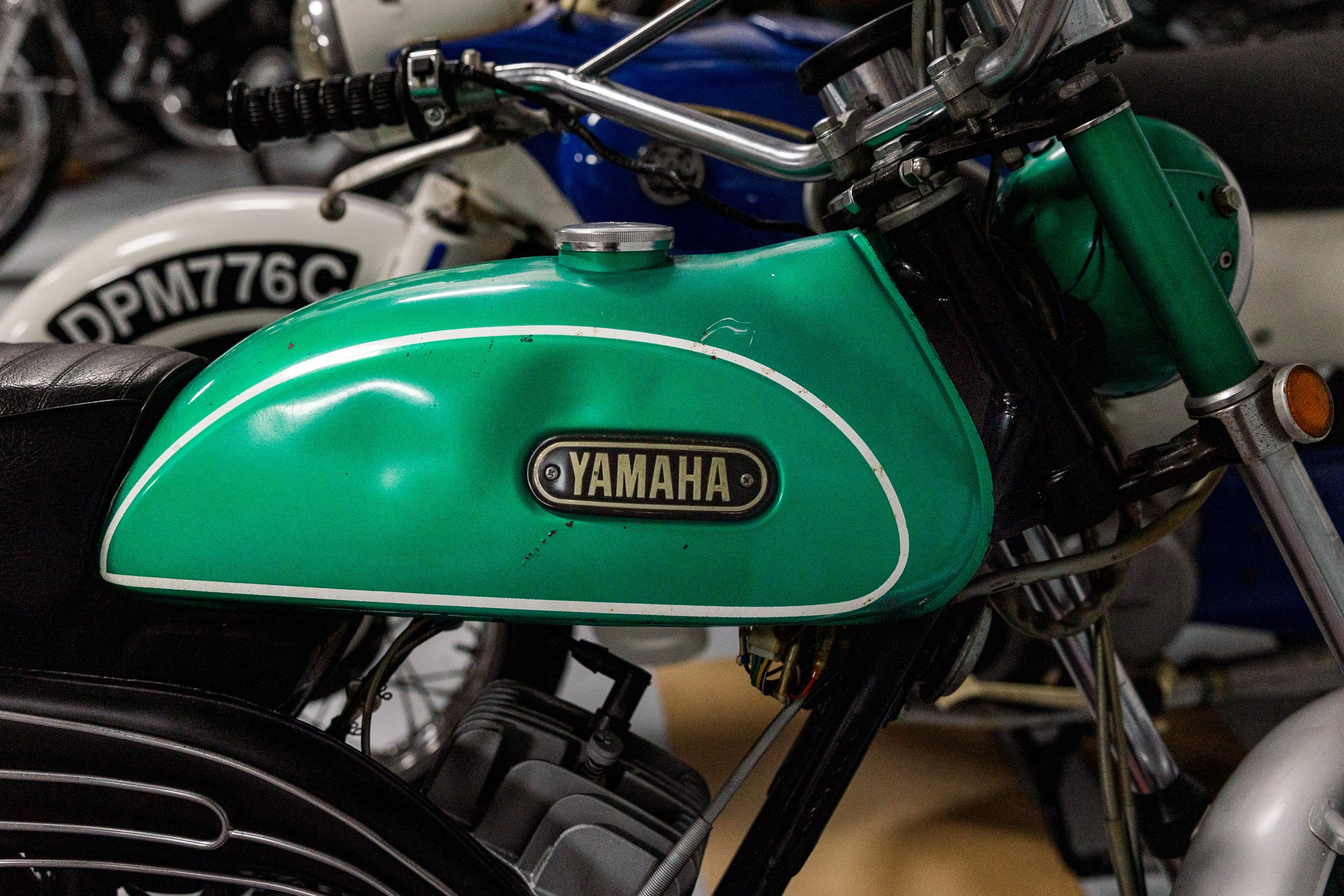 Close-up of a vintage green Yamaha motorcycle dirt bike with black seat and exposed handlebars, part of other motorcycles are visible in the background. Vintage motorcycle collection on display at Birmingham Vintage Cycle in Birmingham Alabama