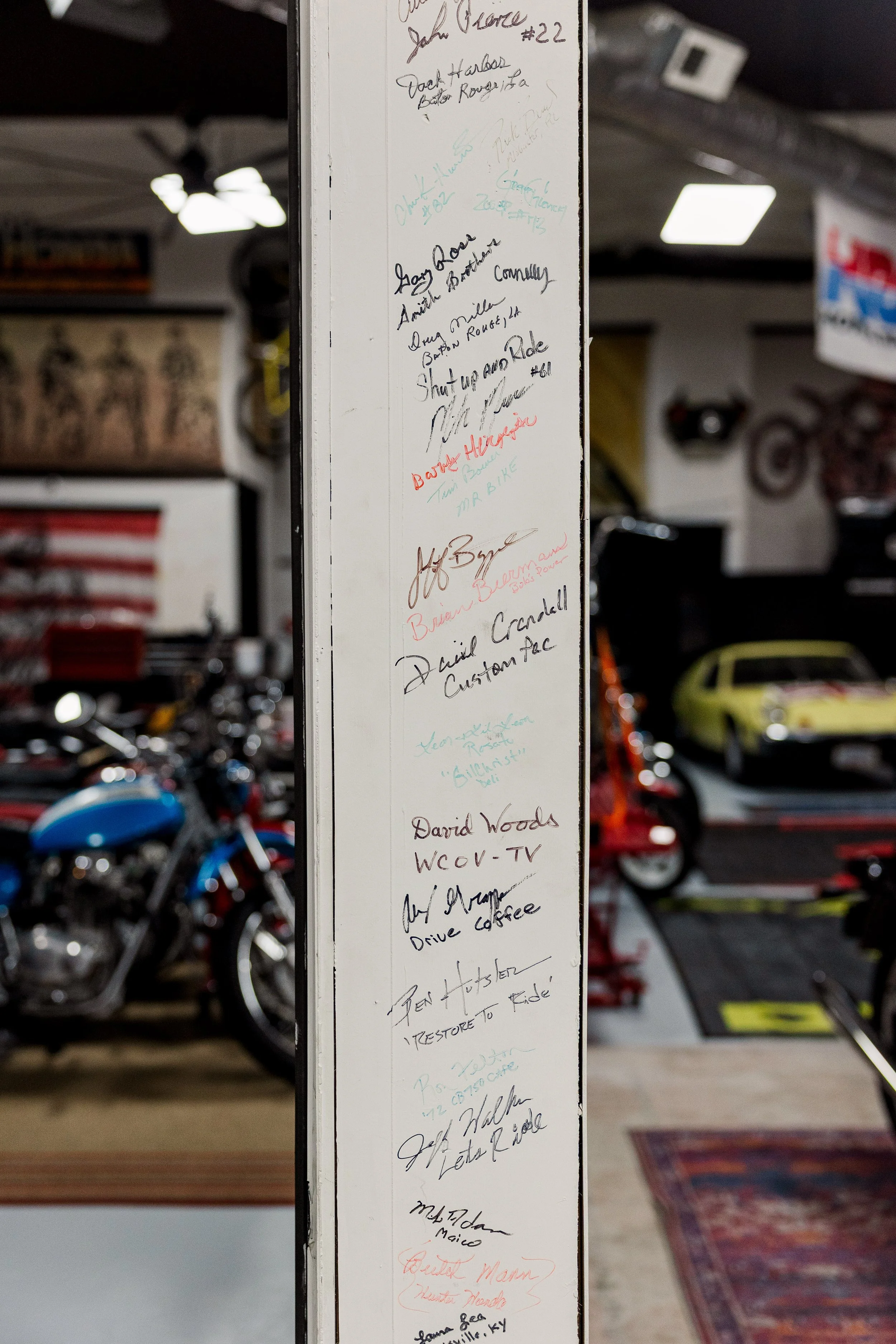 A vertical whiteboard with various signatures and notes written in different colored markers, located inside a garage with cars, motorcycles, and a patterned rug in the background. Inside Birmingham Vintage Cycle, a vintage motorcycle shop