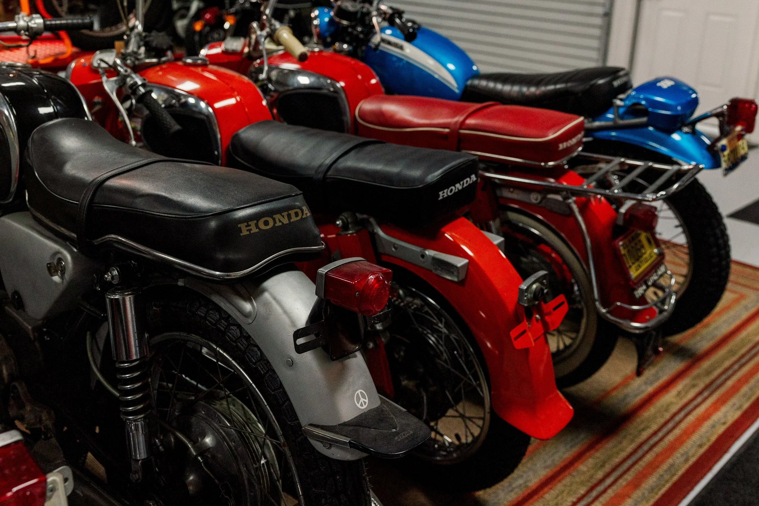 Multiple vintage Honda motorcycles lined up indoors, showing different colors including black, red, grey, and blue, with leather seats and chrome details. Vintage motorcycle collection on display at Birmingham Vintage Cycle in Birmingham Alabama