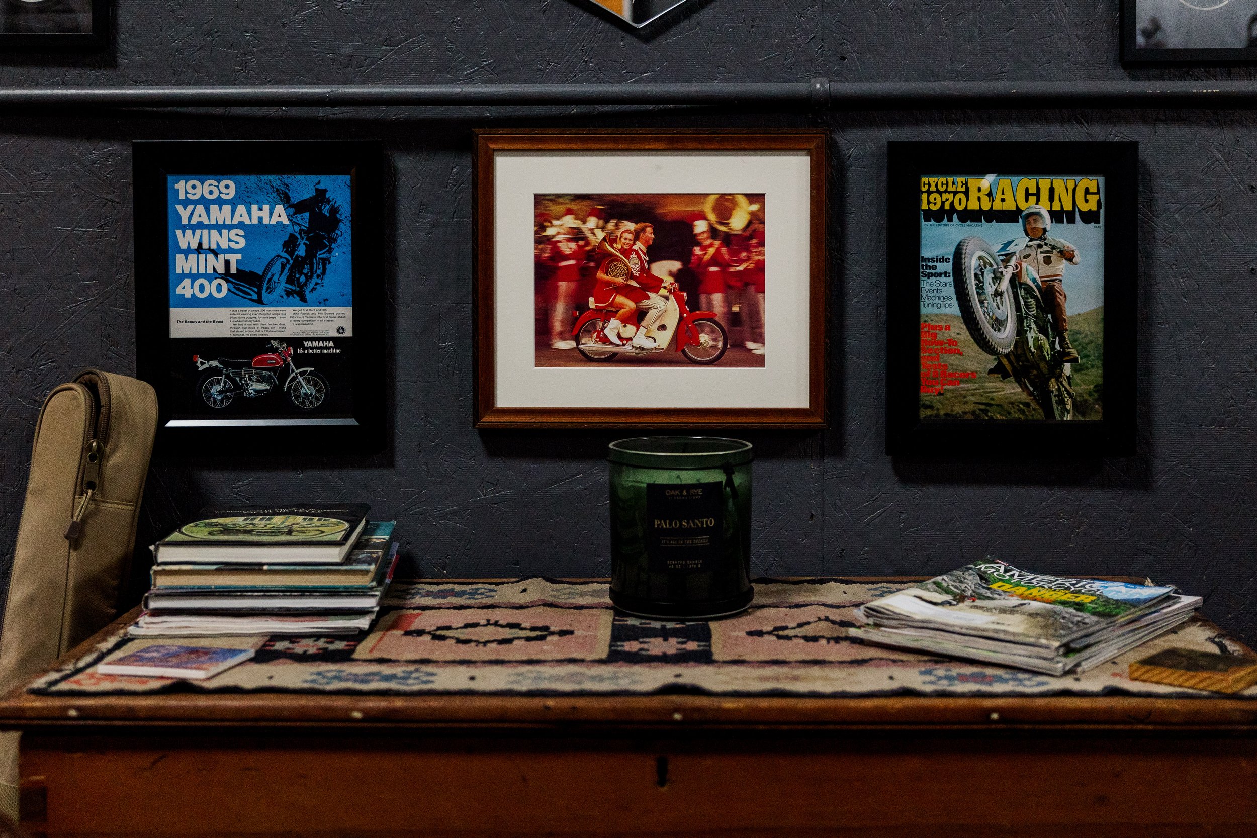 A wall featuring framed vintage motorcycle posters, with a table below displaying a stack of magazines and a small bag, and a book labeled 'Palo Santo' placed on the table. Inside Birmingham Vintage Cycle, a vintage motorcycle shop and community