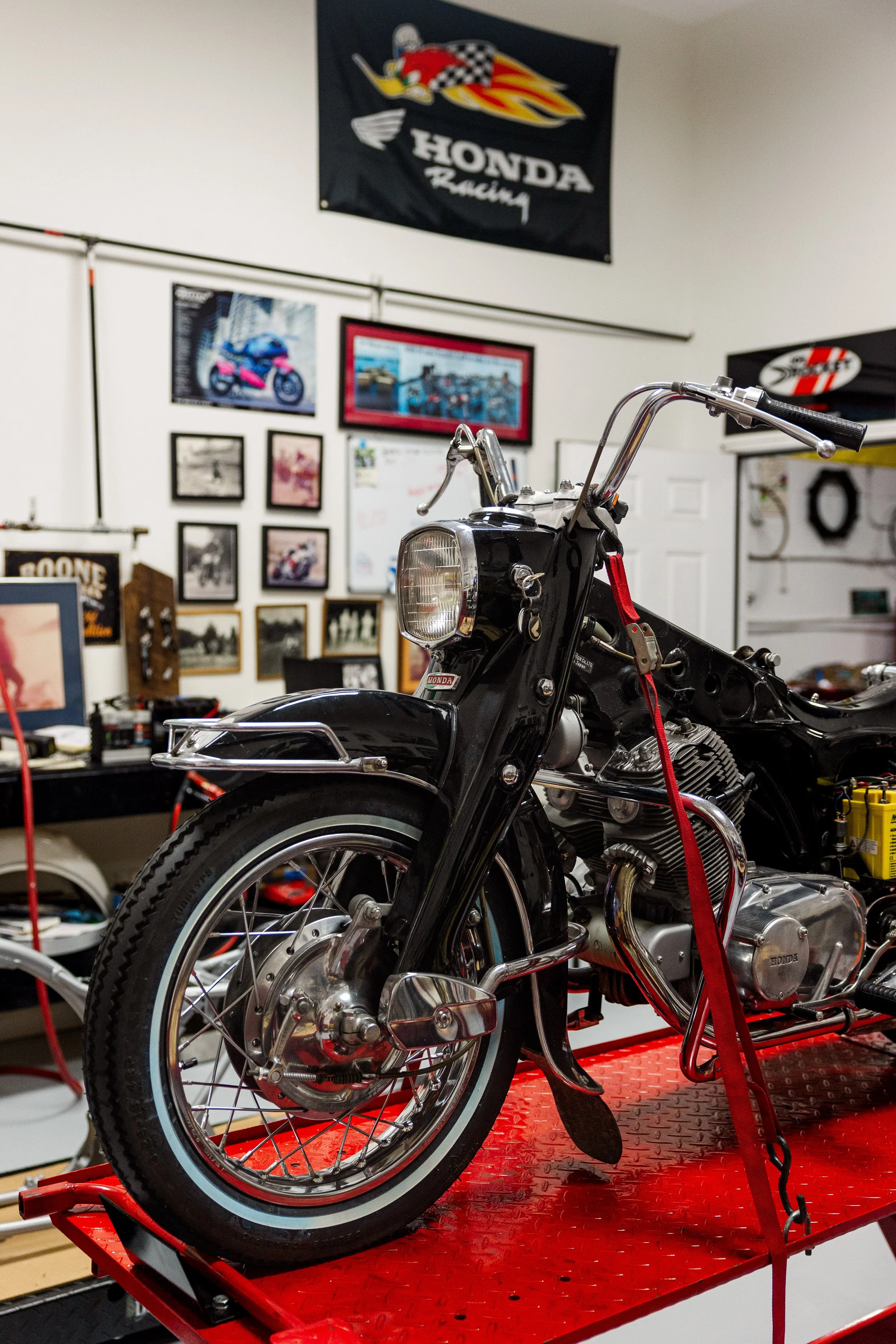 A black vintage motorcycle on a red motorcycle lift inside a garage, with Honda Racing banners and framed pictures of motorcycles on the wall in the background. Vintage motorcycle detail at Birmingham Vintage Cycle, a vintage motorcycle restoration 