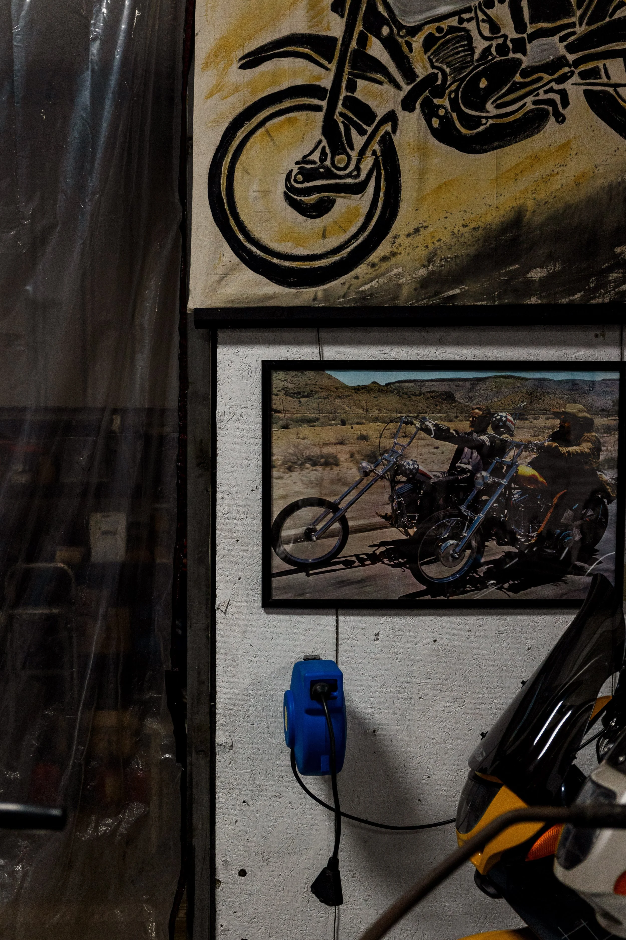 A framed photograph on the wall showing two bikers riding mountain bikes in a desert landscape with mountains in the background. Above the photograph, a large painting depicts part of a motorcycle wheel and a rider's legs.