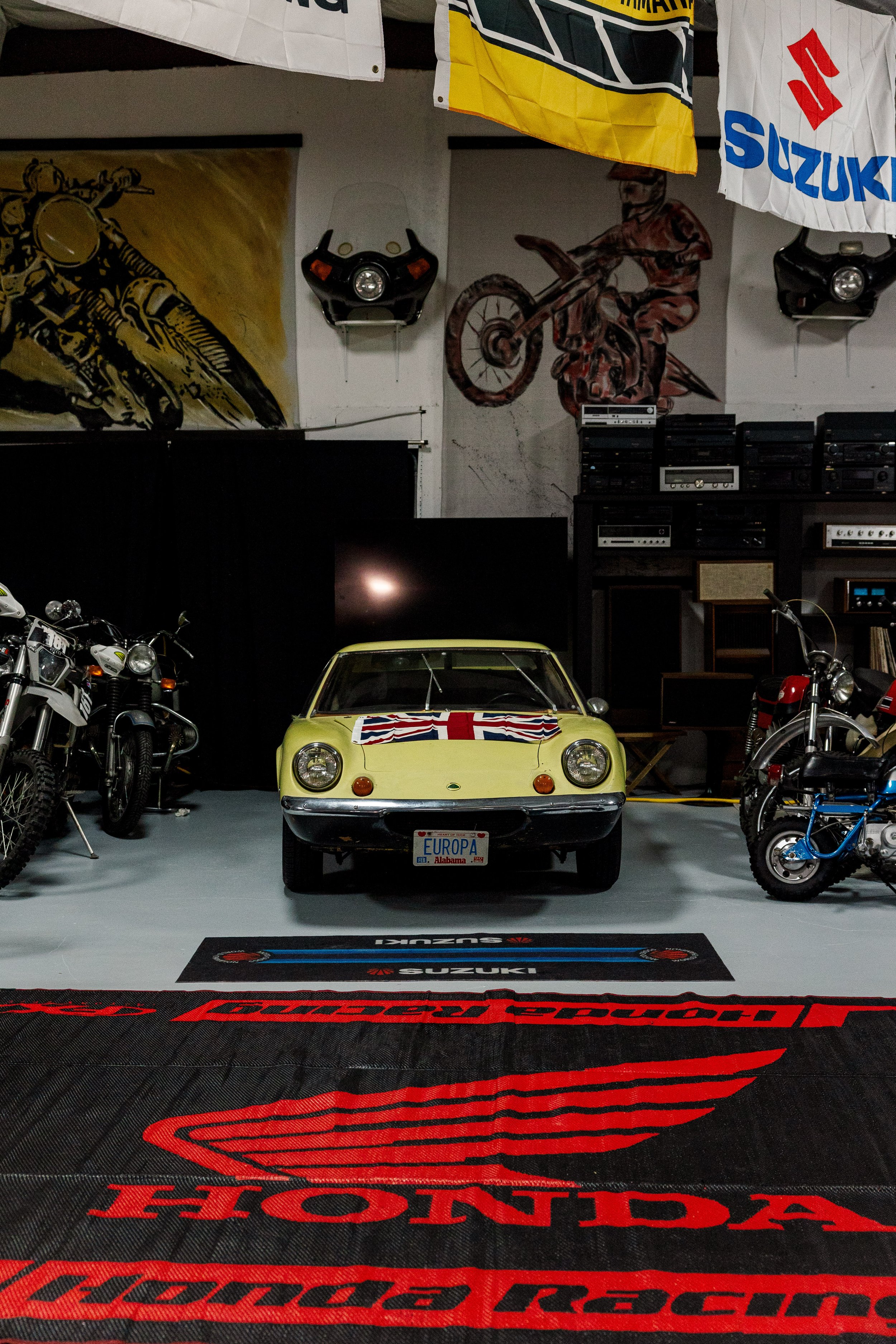 A yellow vintage Lotus car with a Union Jack flag painted on the front hood, parked in a garage. The garage walls display motorcycle artwork, banners, and flags with motorcycle and Suzuki logos. Various motorcycles and motorcycle parts are also visib