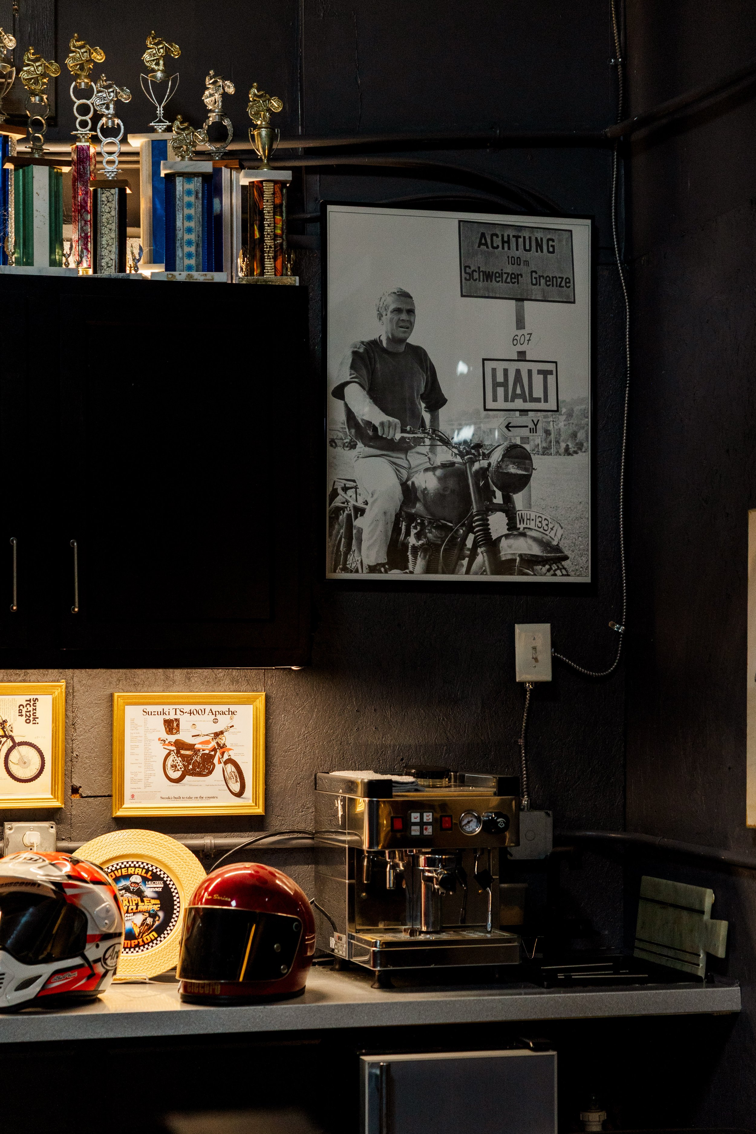 A collection of motorcycle helmets, trophies, framed pictures, and a coffee machine on a dark-colored counter in a room. The framed pictures show motorcycles, and one features a man on a motorcycle with signs in German and English. Inside BVC 