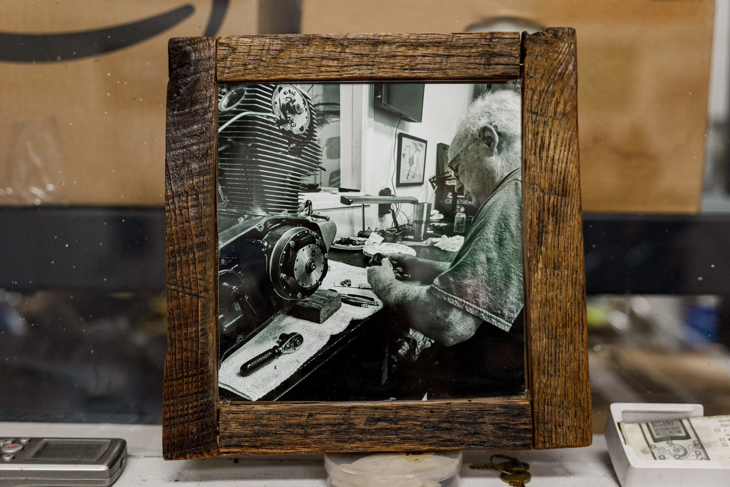 A framed black and white photo of an elderly man working on a mechanical project, possibly a motorcycle engine, sitting at a workbench cluttered with tools.