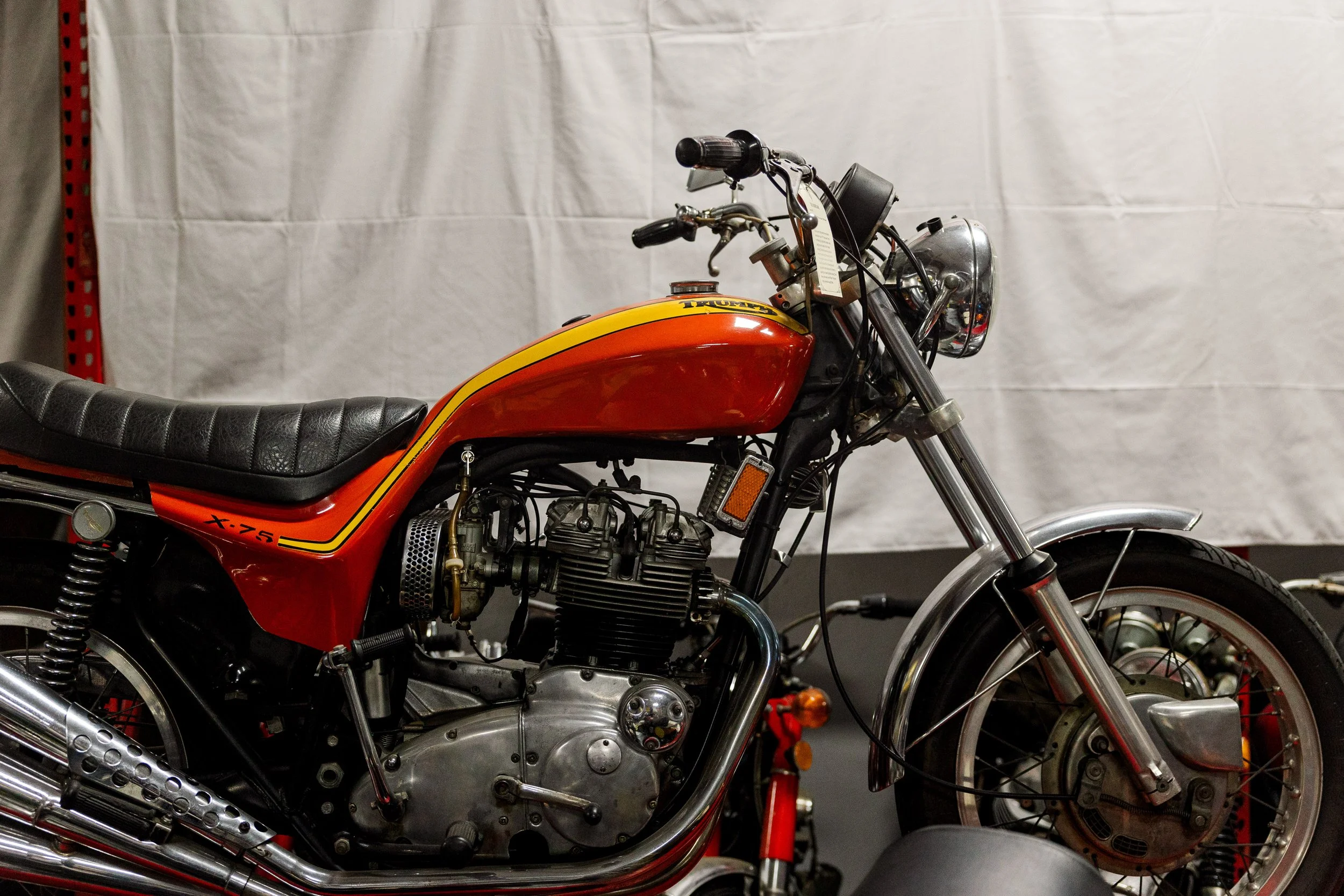 A vintage Triumph motorcycle with a red and yellow tank, black seat, and chrome details, parked indoors against a white backdrop. Restored vintage motorcycle in the Birmingham Vintage Cycle collection in Birmingham Alabama