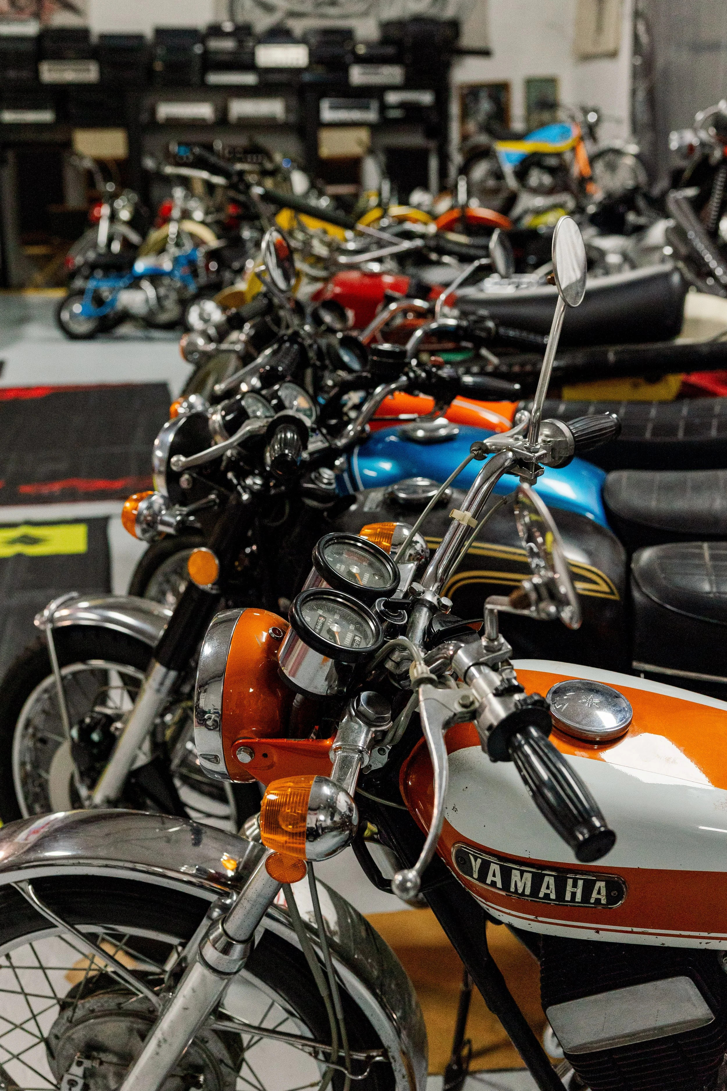 Several vintage motorcycles of various colors including orange, black, yellow, and blue, parked indoors, with a focus on an orange Yamaha motorcycle in the foreground. Vintage motorcycle collection on display at Birmingham Vintage Cycle in Birmingham