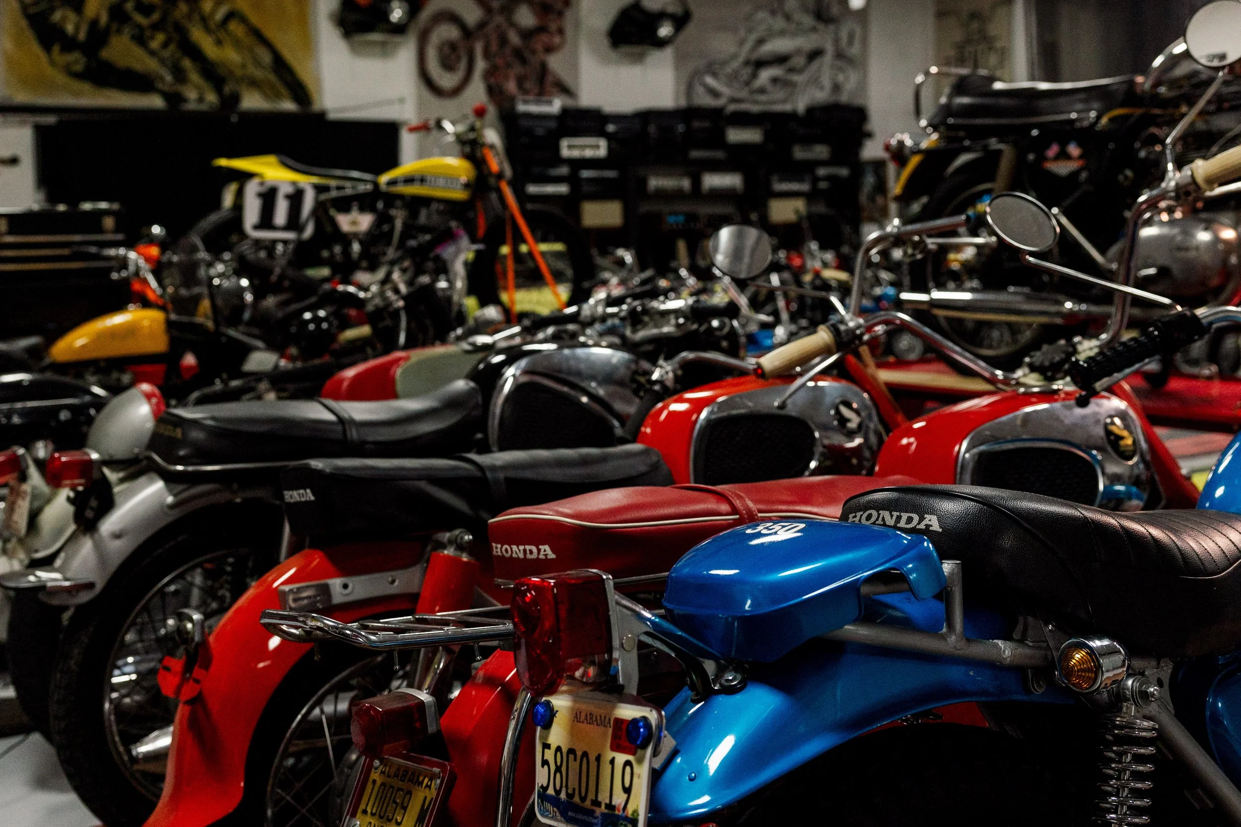Multiple vintage Honda motorcycles parked indoors, some with Alabama license plates, in a dimly lit room with artwork on the walls. Vintage motorcycle collection on display at Birmingham Vintage Cycle in Birmingham Alabama
