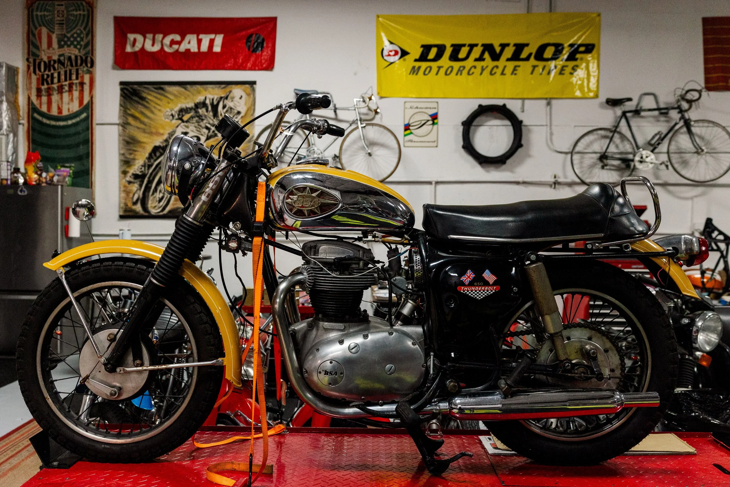 A vintage motorcycle with a yellow front fender and black body, parked on a red platform inside a garage with bikes and posters on the wall. Vintage motorcycle collection on display at Birmingham Vintage Cycle in Birmingham Alabama