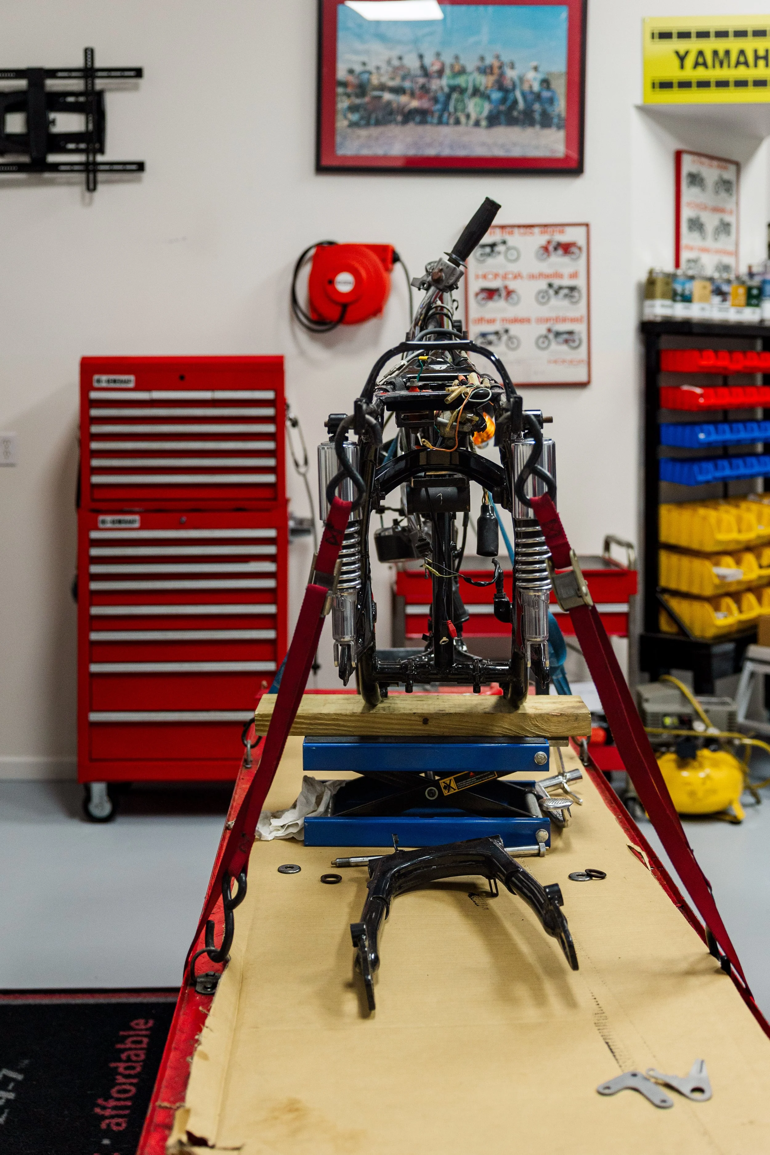 A motorcycle frame in the process of assembly or repair is set on a workbench in a workshop. Tools and small parts are scattered around, with a red tool chest and various posters and storage bins visible in the background.