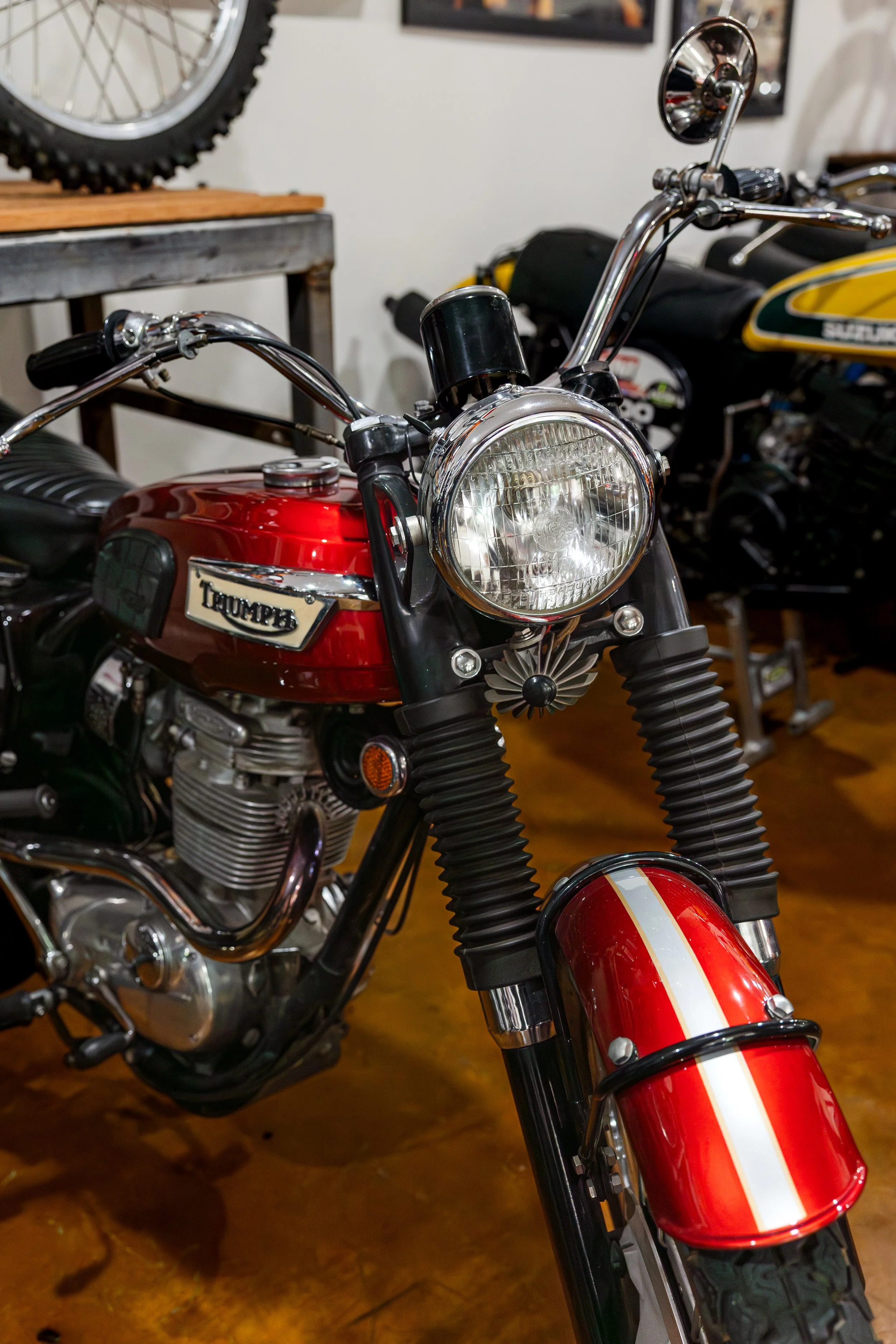 Close-up of a vintage red Triumph motorcycle's front end, including the headlight, handlebars, and front fork with a red and white striped fender, in a room with other motorcycles and a workbench in the background.