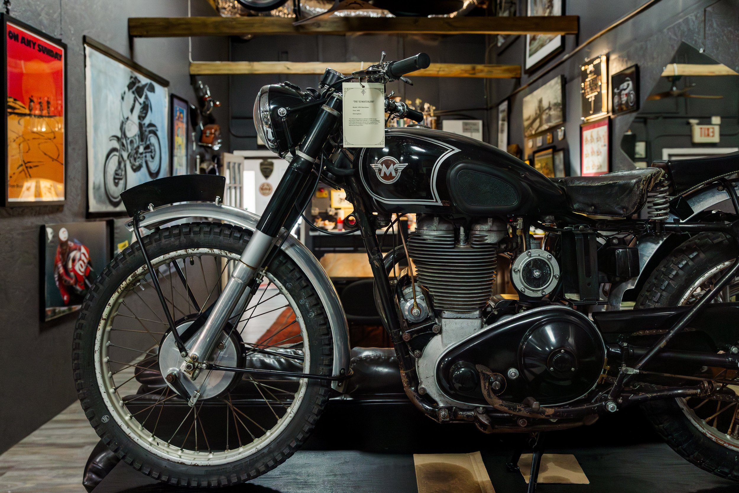 A vintage black motorcycle with a white emblem on the gas tank, displayed indoors with art and posters on the walls, and a tag hanging from the handlebars. Vintage motorcycle collection on display at Birmingham Vintage Cycle in Birmingham Alabama