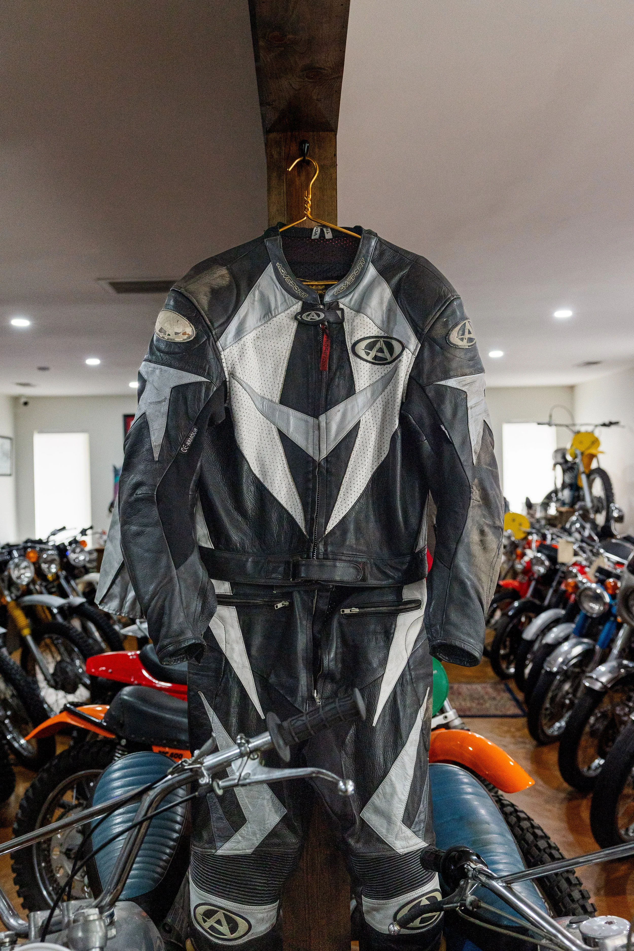 A motorcycle jacket hanging on a yellow hanger from a wooden ceiling beam in a motorcycle showroom with several bikes displayed in the background. Vintage motorcycle collection on display at Birmingham Vintage Cycle in Birmingham Alabama