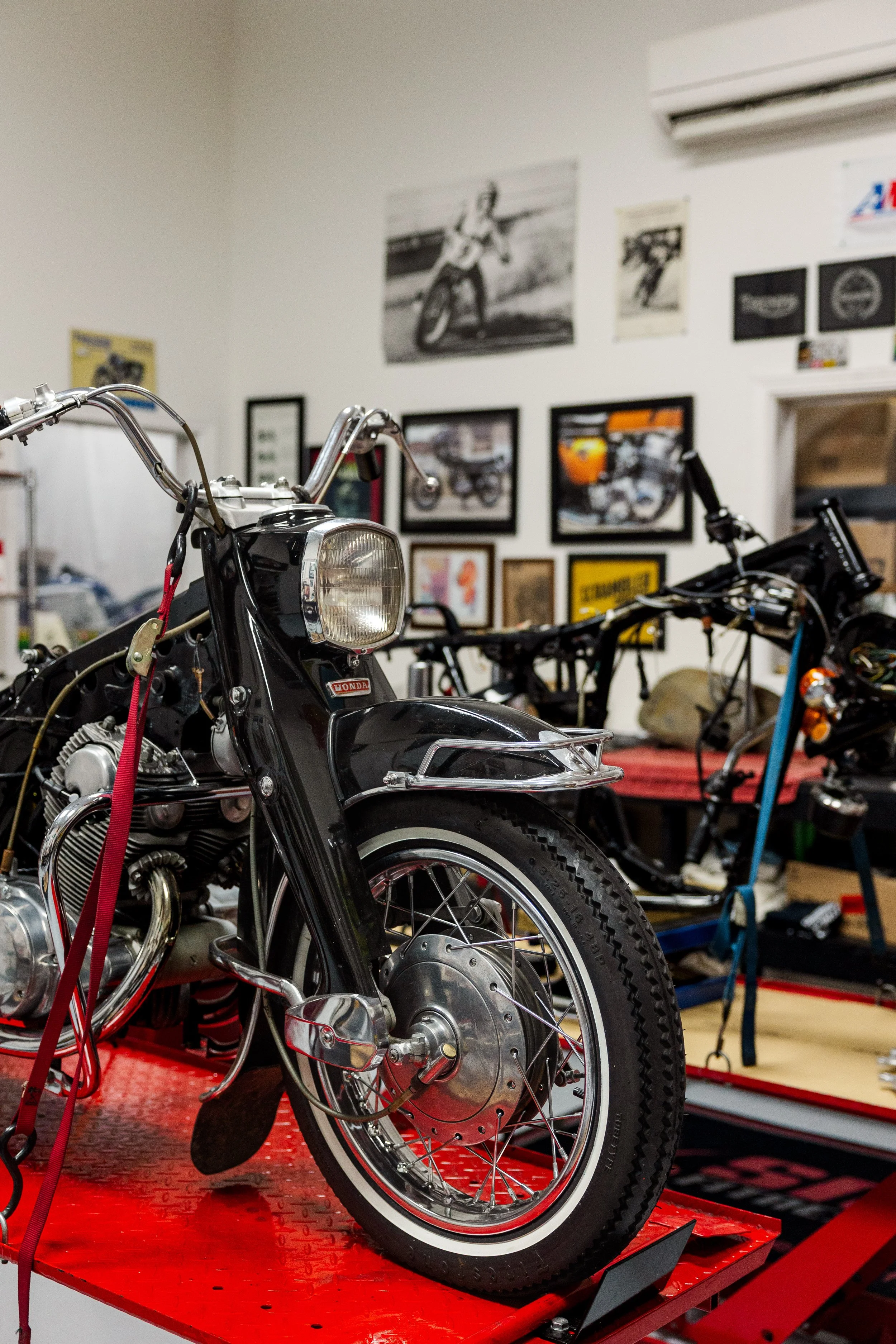 A vintage motorcycle on a red workbench in a garage, with framed posters and photos of motorcycles on the wall behind it. Vintage motorcycle detail at Birmingham Vintage Cycle, a vintage motorcycle restoration shop in Birmingham Alabama