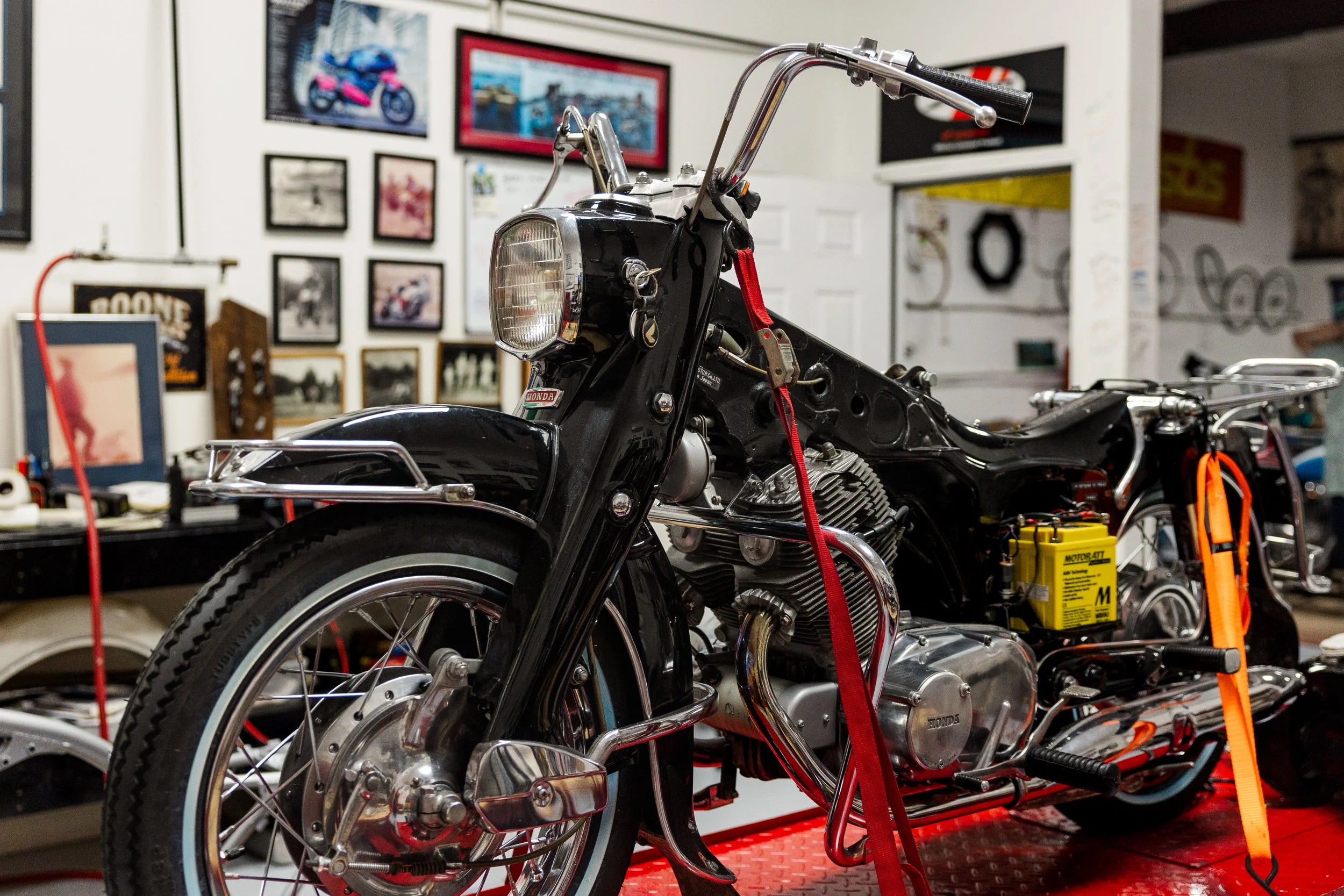 A black vintage Honda motorcycle on a red platform in a workshop, surrounded by tools and framed pictures on the wall. Vintage motorcycle detail at Birmingham Vintage Cycle, a vintage motorcycle restoration shop in Birmingham Alabama