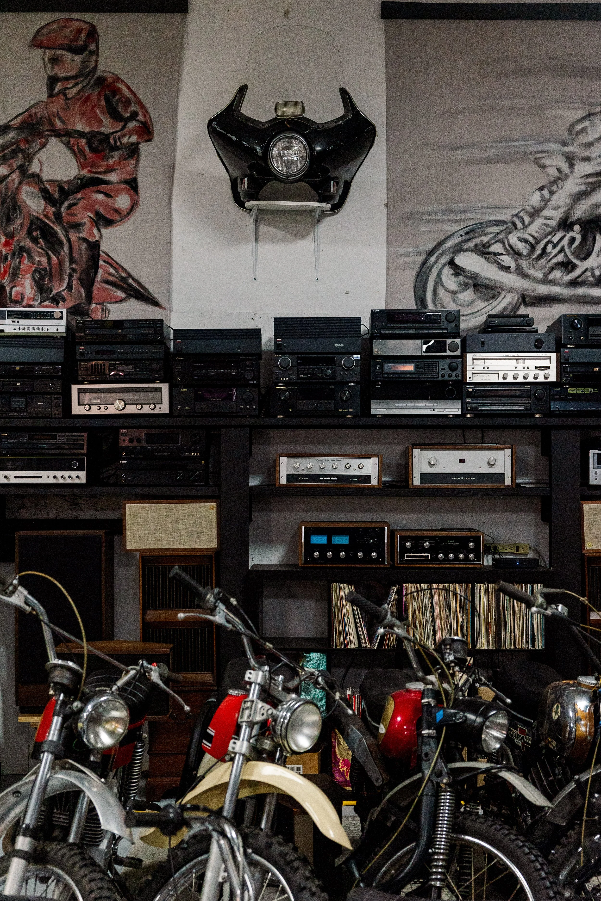 A collection of vintage motorcycles parked in front of a black shelving unit containing vinyl records, with a large mural of a cyclist on the wall behind. Vintage motorcycle collection on display at Birmingham Vintage Cycle in Birmingham Alabama